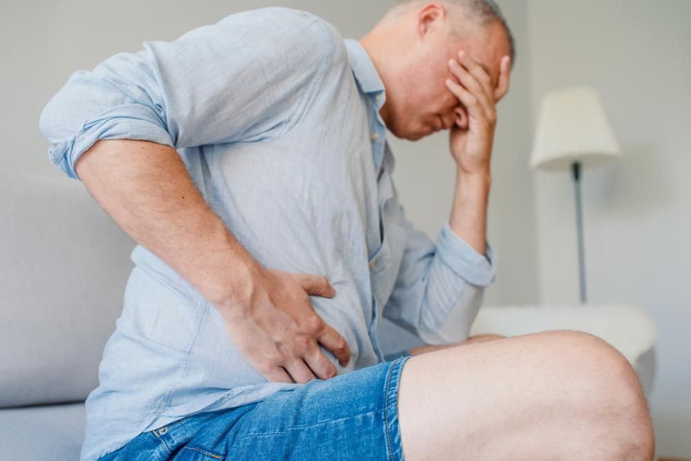 A Man Is Sitting on A Couch Holding His Stomach in Pain — Action Physiotherapy in Cooks Hill, NSW