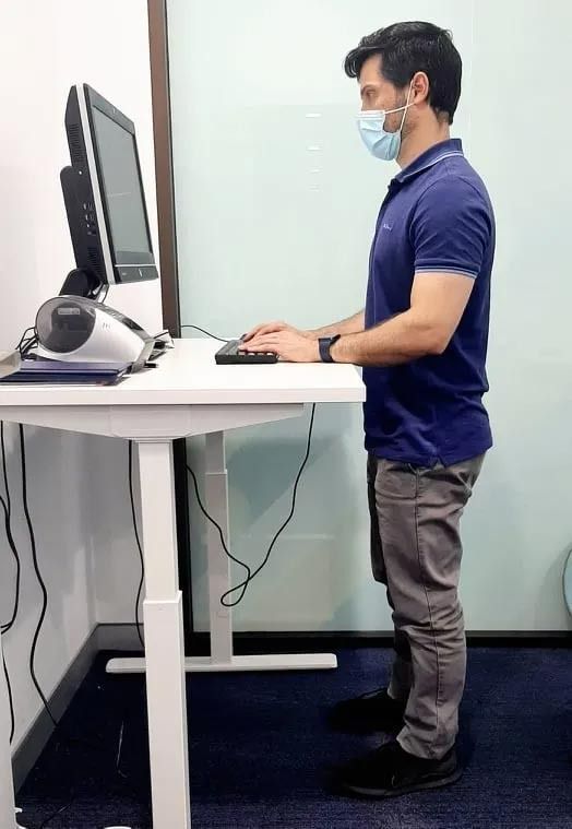 A Man Wearing a Mask Is Standing at A Standing Desk Using a Computer — Action Physiotherapy in Cooks Hill, NSW