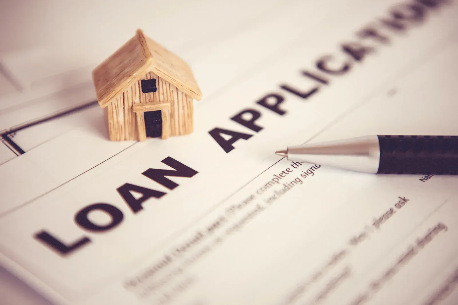 A small wooden house model sits on a loan application document next to a black ballpoint pen.