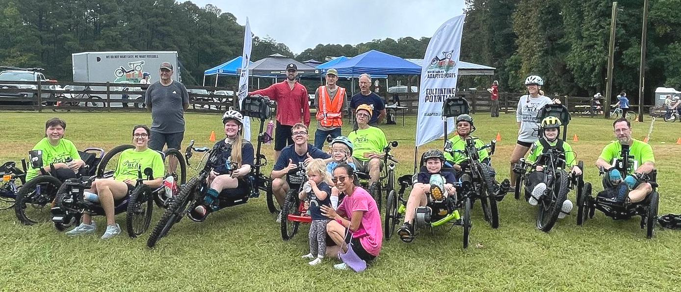 Group of adapted cyclists posing with their handcycles and recumbents on a grassy field near tents and trees at an Kanata Cross