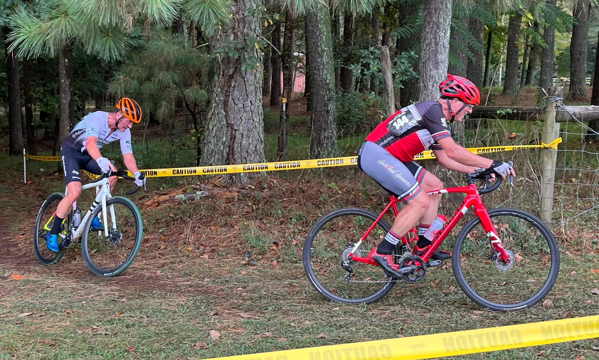 Two cyclists ride on a muddy forest trail, one in red leading and one in gray trailing behind.