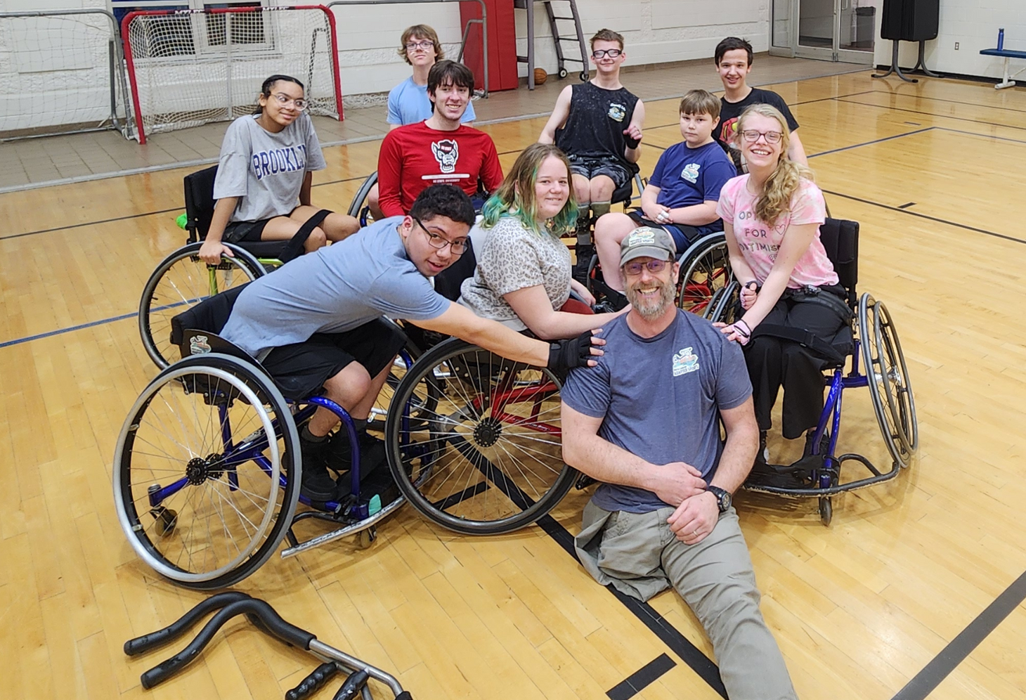 Group of kids and young adults in sport wheelchairs posing and smiling in a gym with Coach Wes Hall sitting on floor