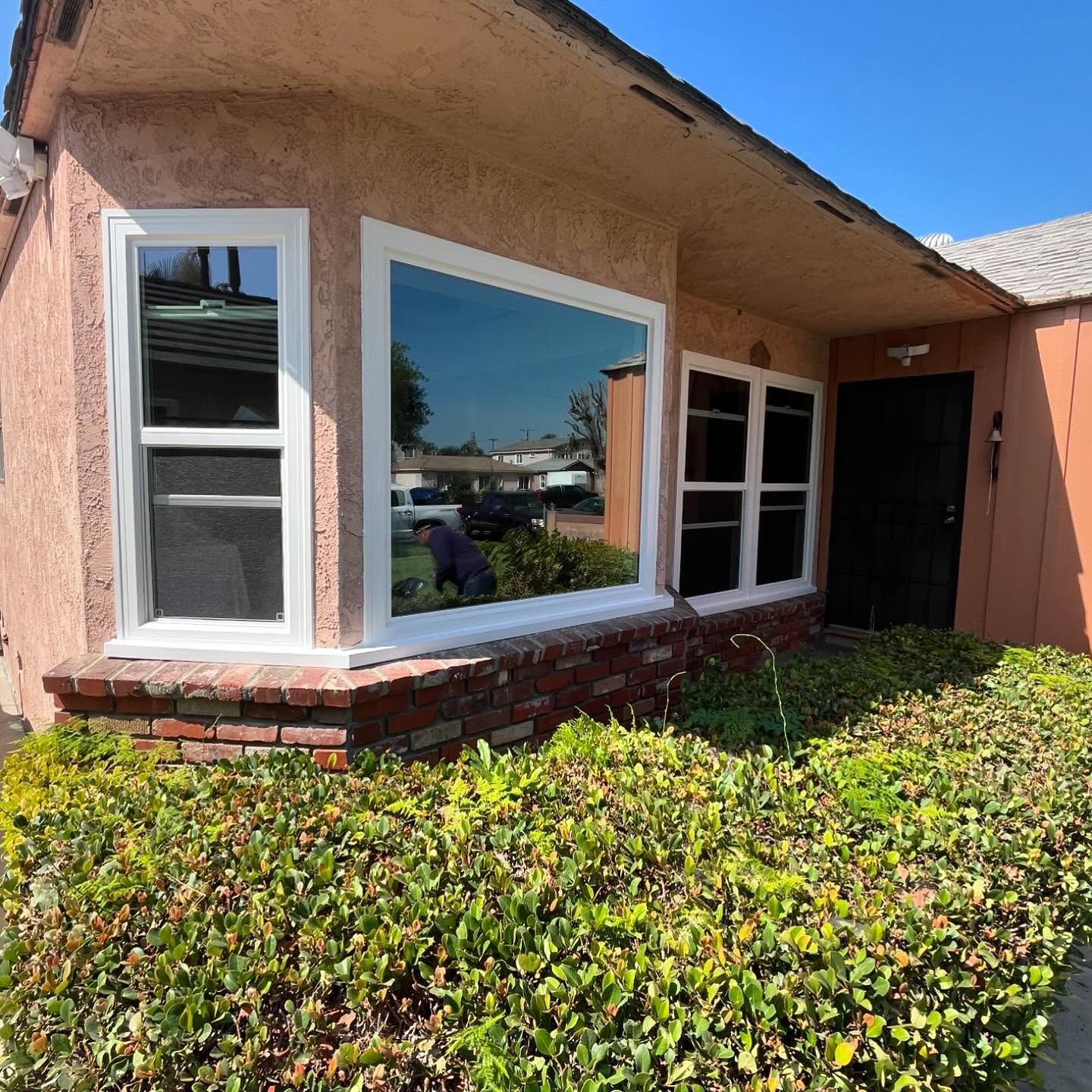 Front of a peach stucco house with white-trimmed windows and a green hedge in sunlight