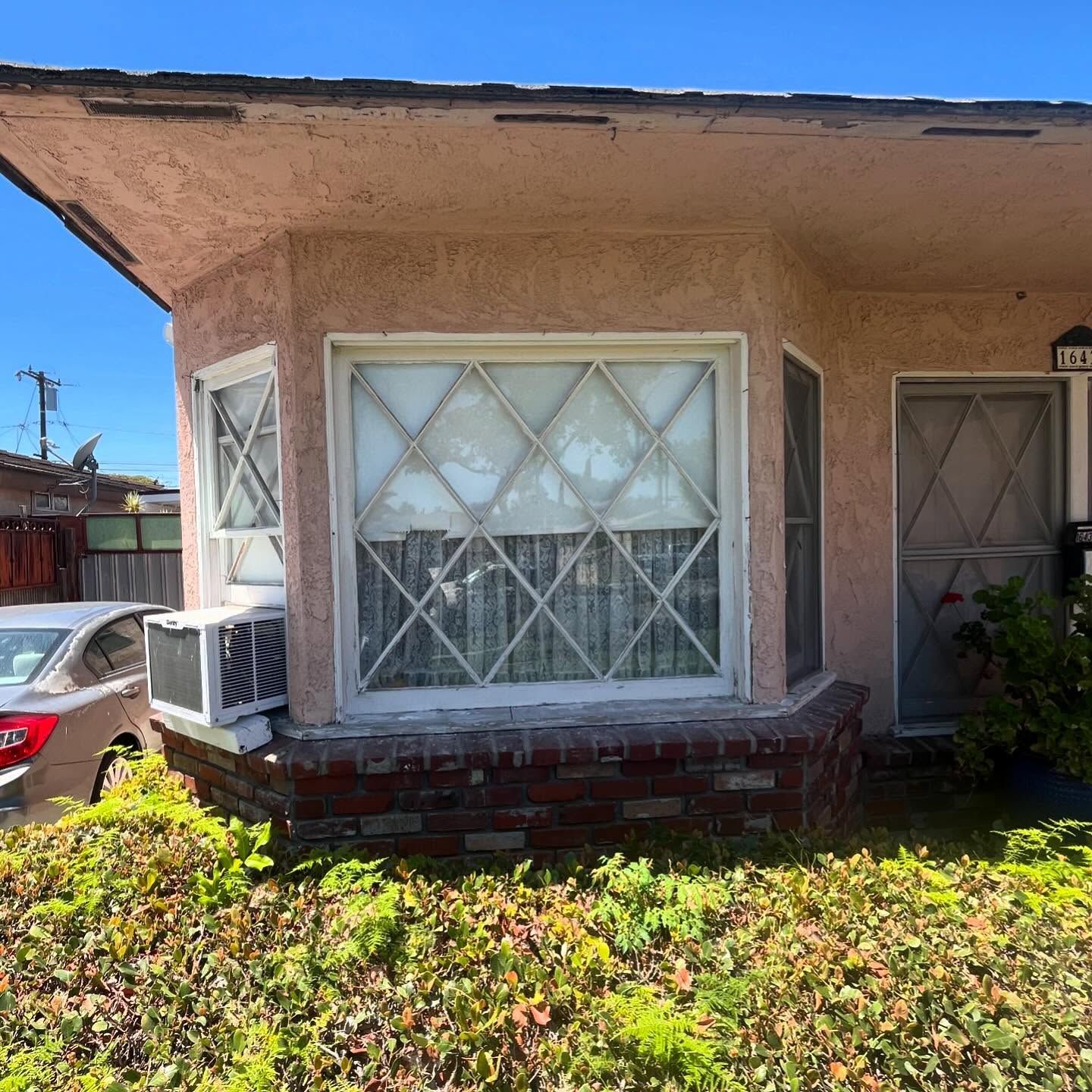 Stucco house front with barred windows, a small porch, and an air conditioner, partly covered by shrubs.