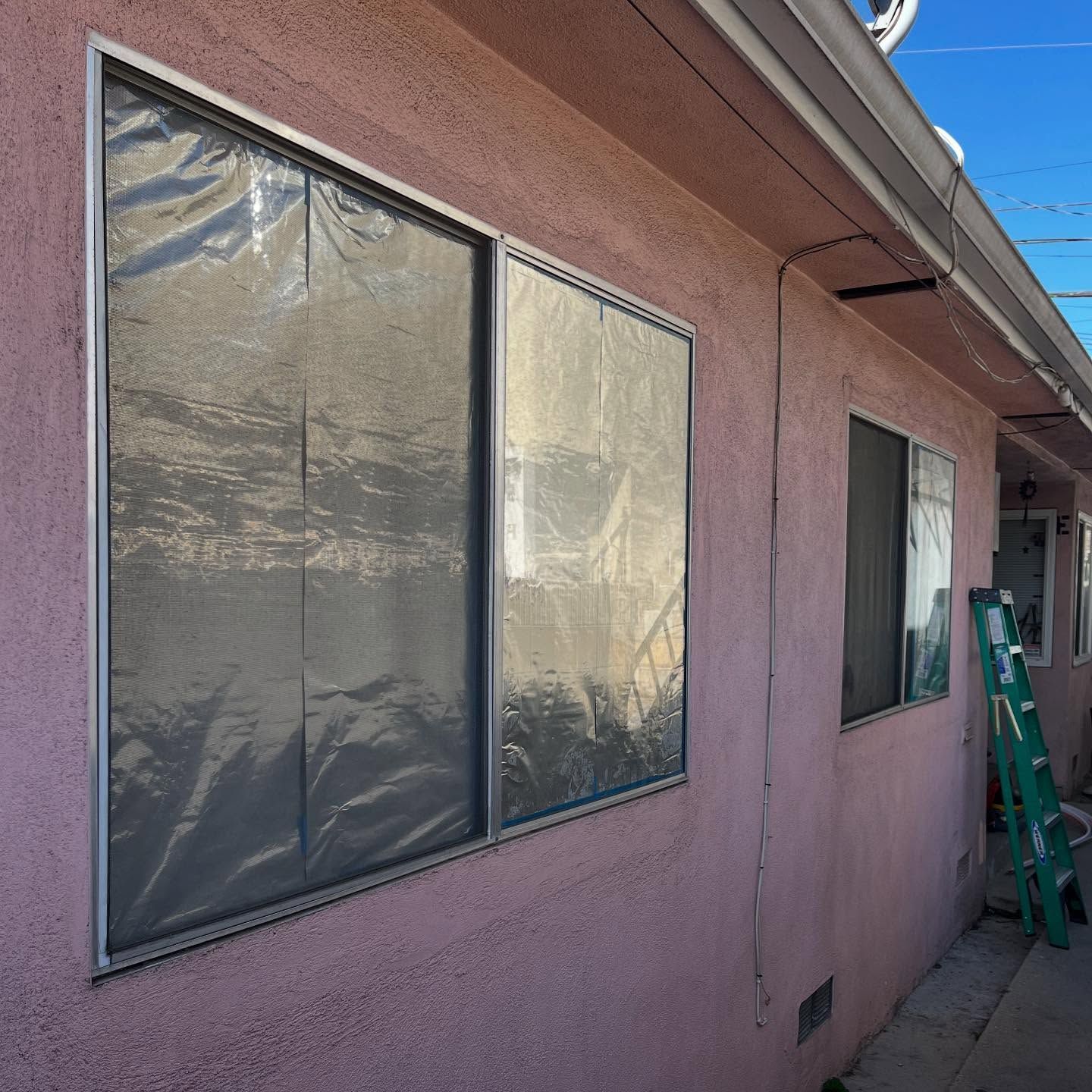 House exterior with pink stucco wall, screened windows, and a green ladder beside a narrow side walkway