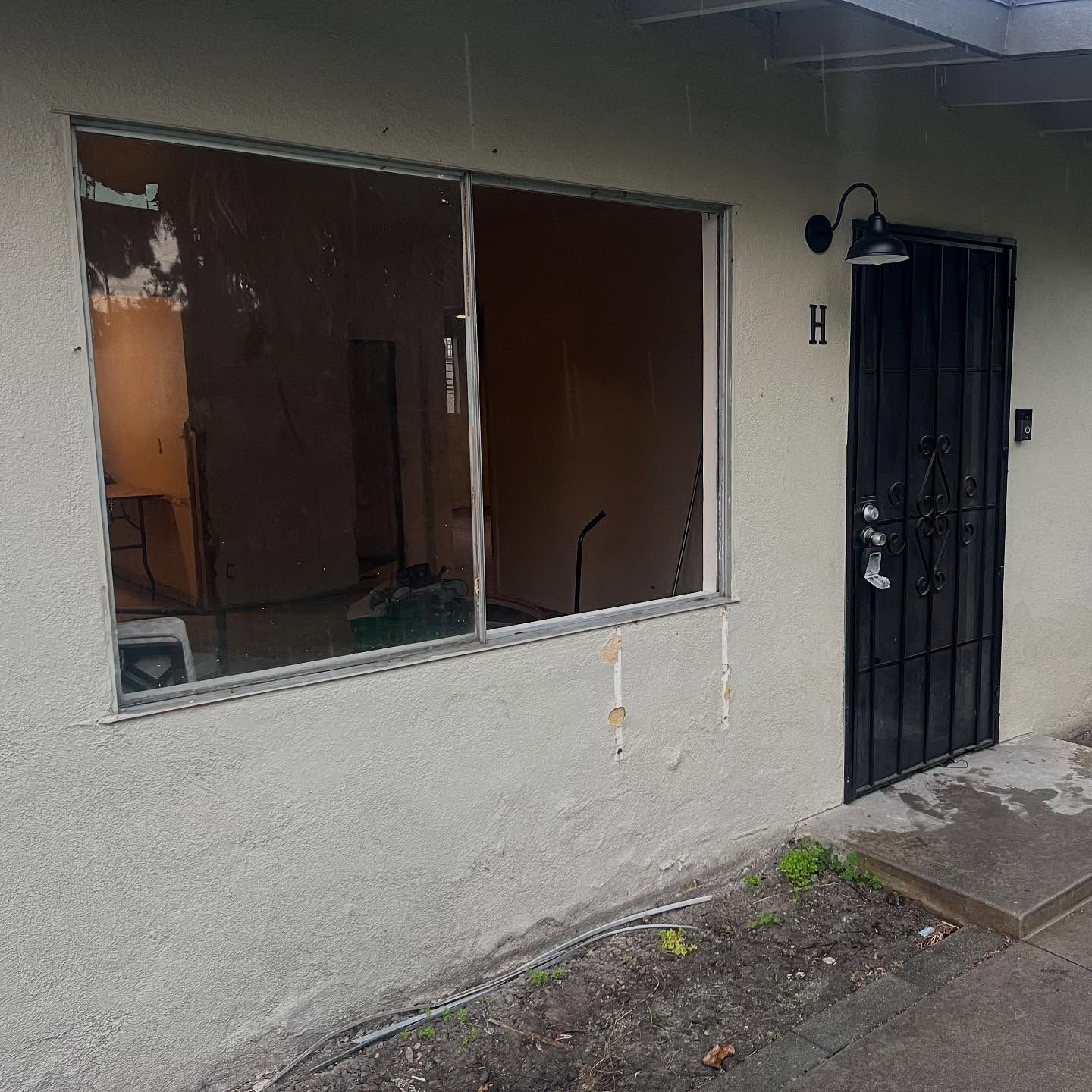 Damaged storefront with broken window, white stucco wall, and black security door beside a sidewalk.