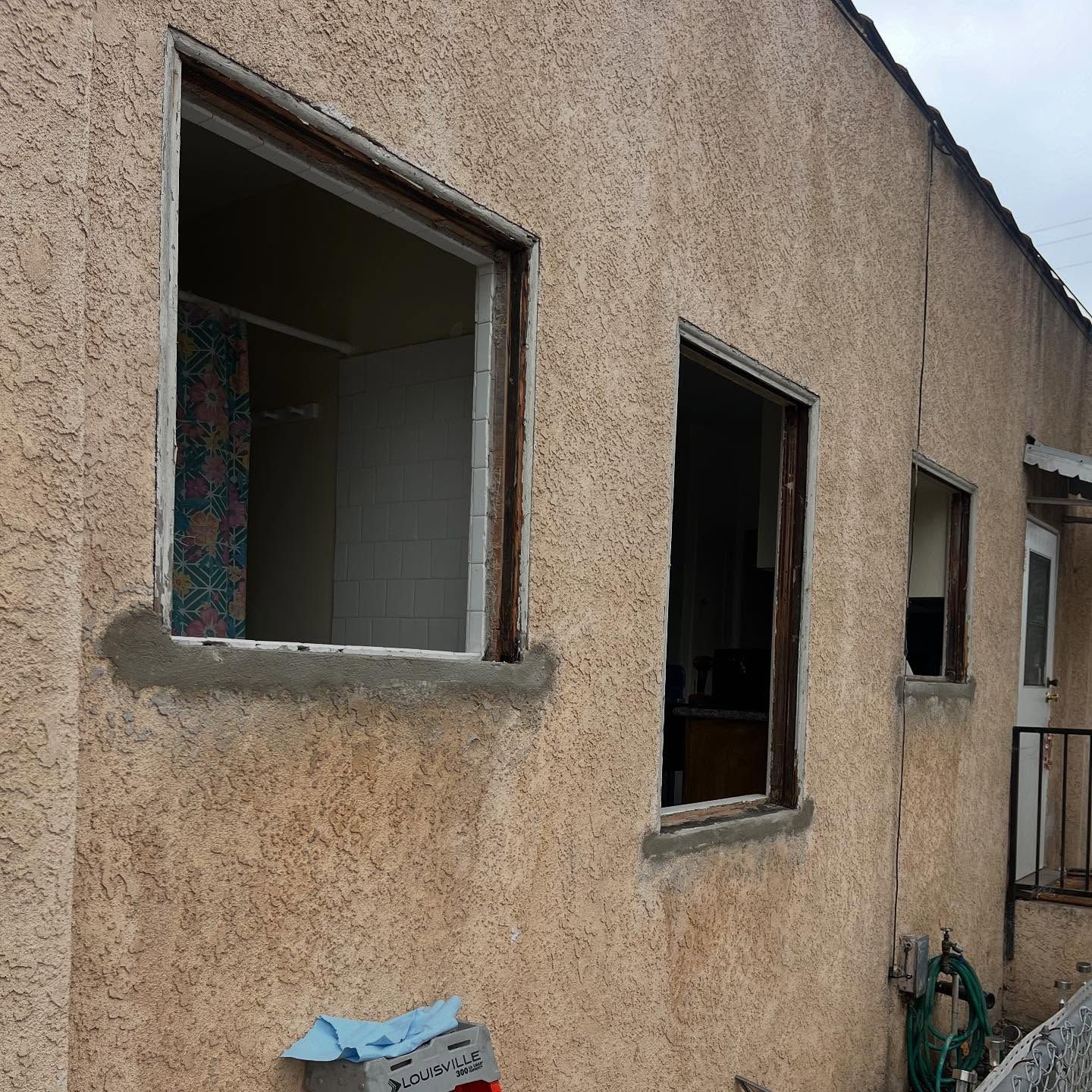 Three open windows in a beige stucco wall beside a green garden hose and broom