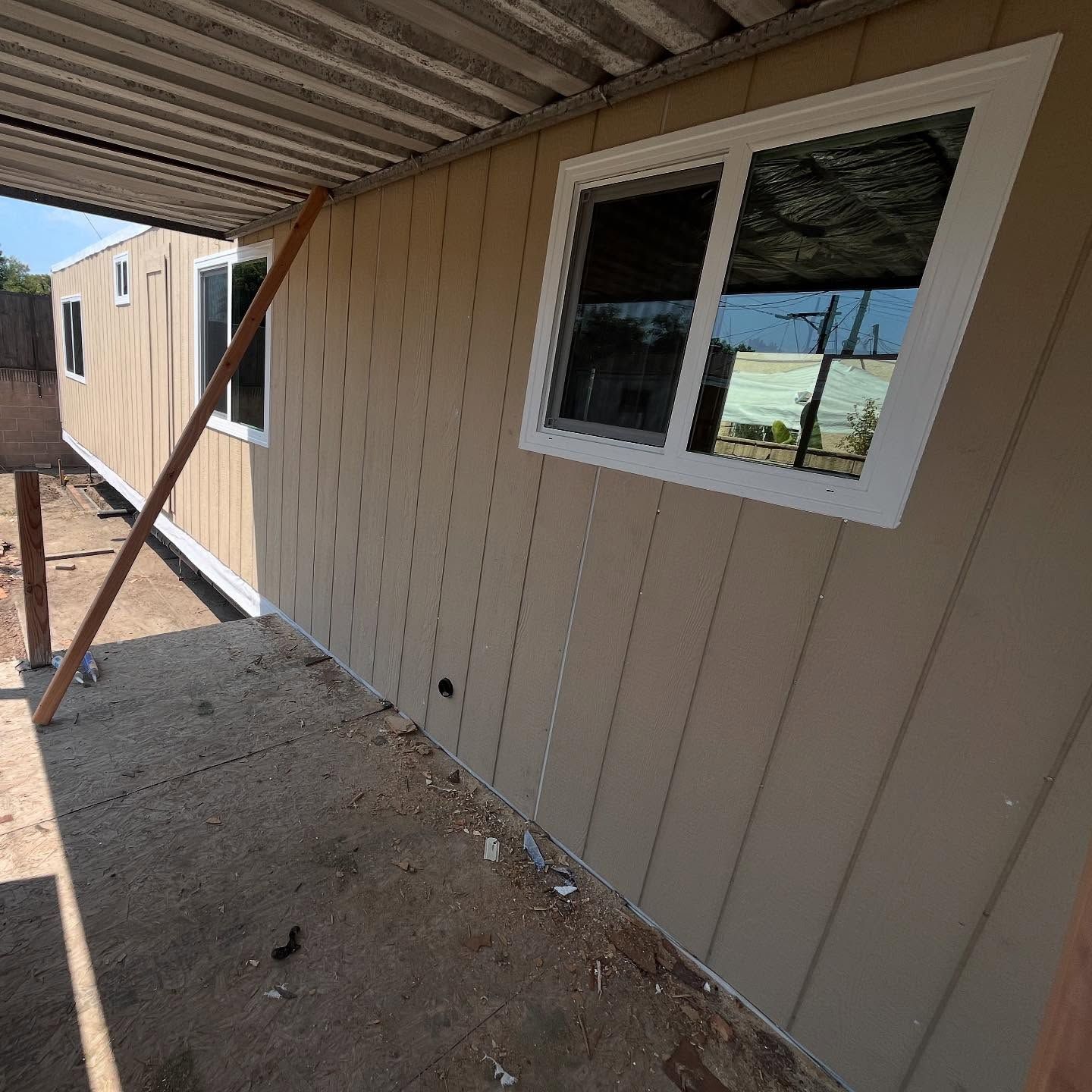 Side of a beige modular home with a white-framed window and covered porch in a sandy yard.