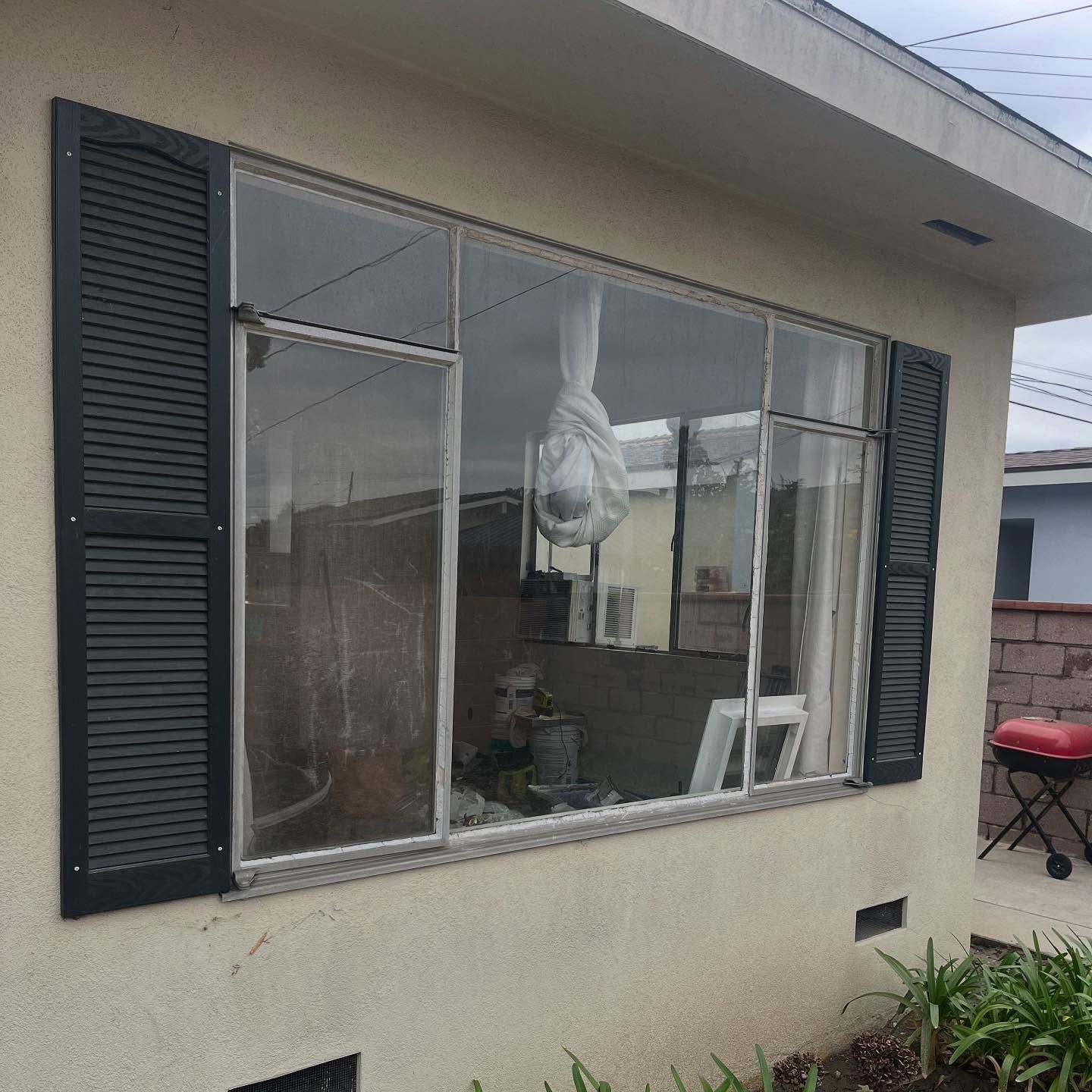 Exterior window with black shutters, a hanging white bag, and a small garden beside a beige house