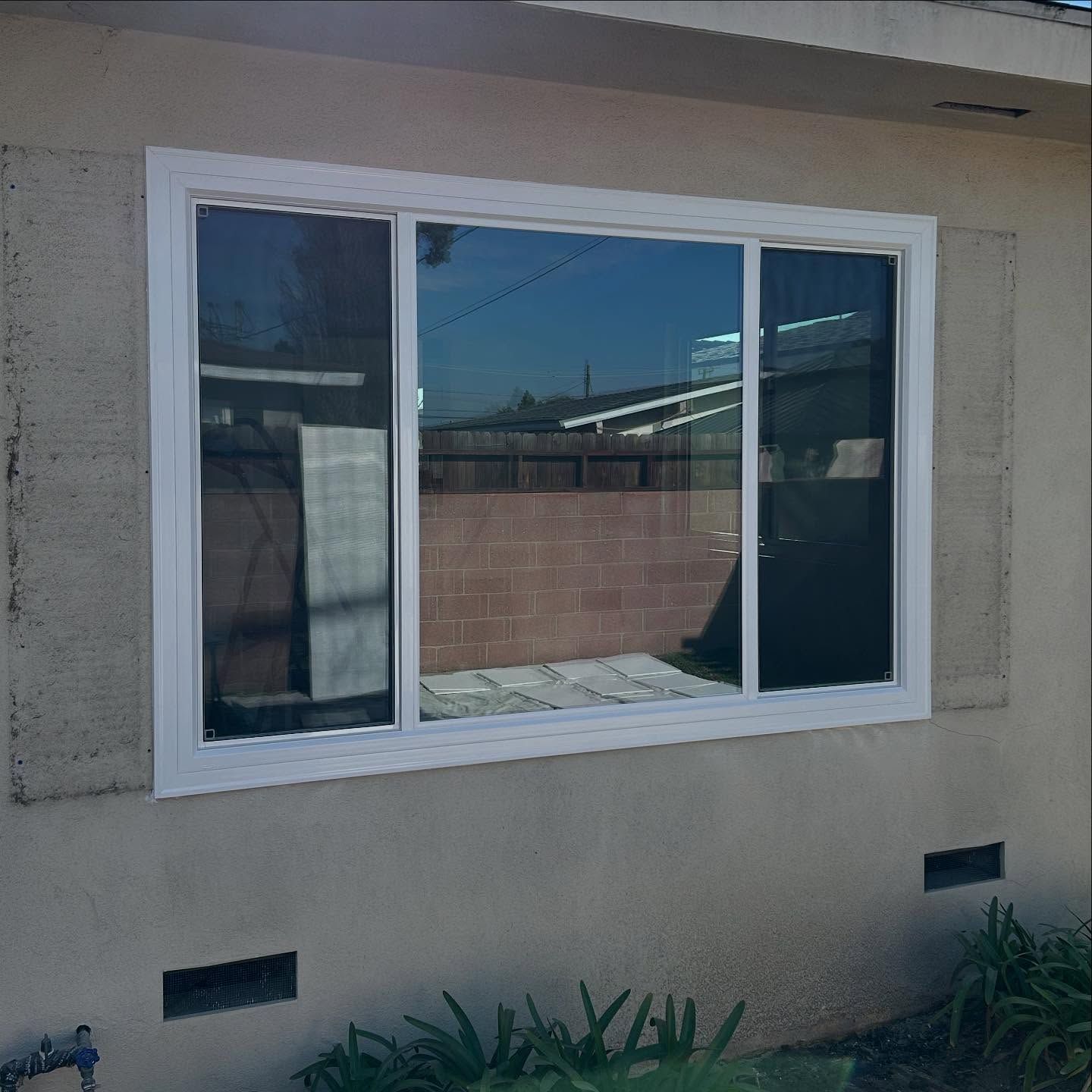 White-framed sliding window on a beige stucco wall with small vents and plants below