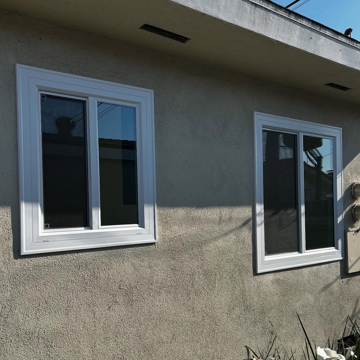 Two white-framed windows on a beige stucco house exterior in sunlight