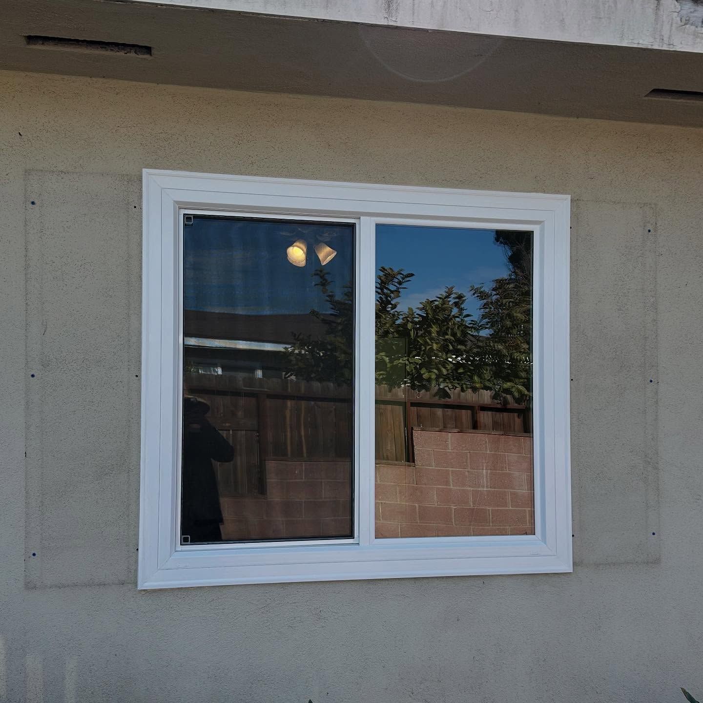 White-framed sliding window set into a beige stucco wall, reflecting a backyard fence and trees.