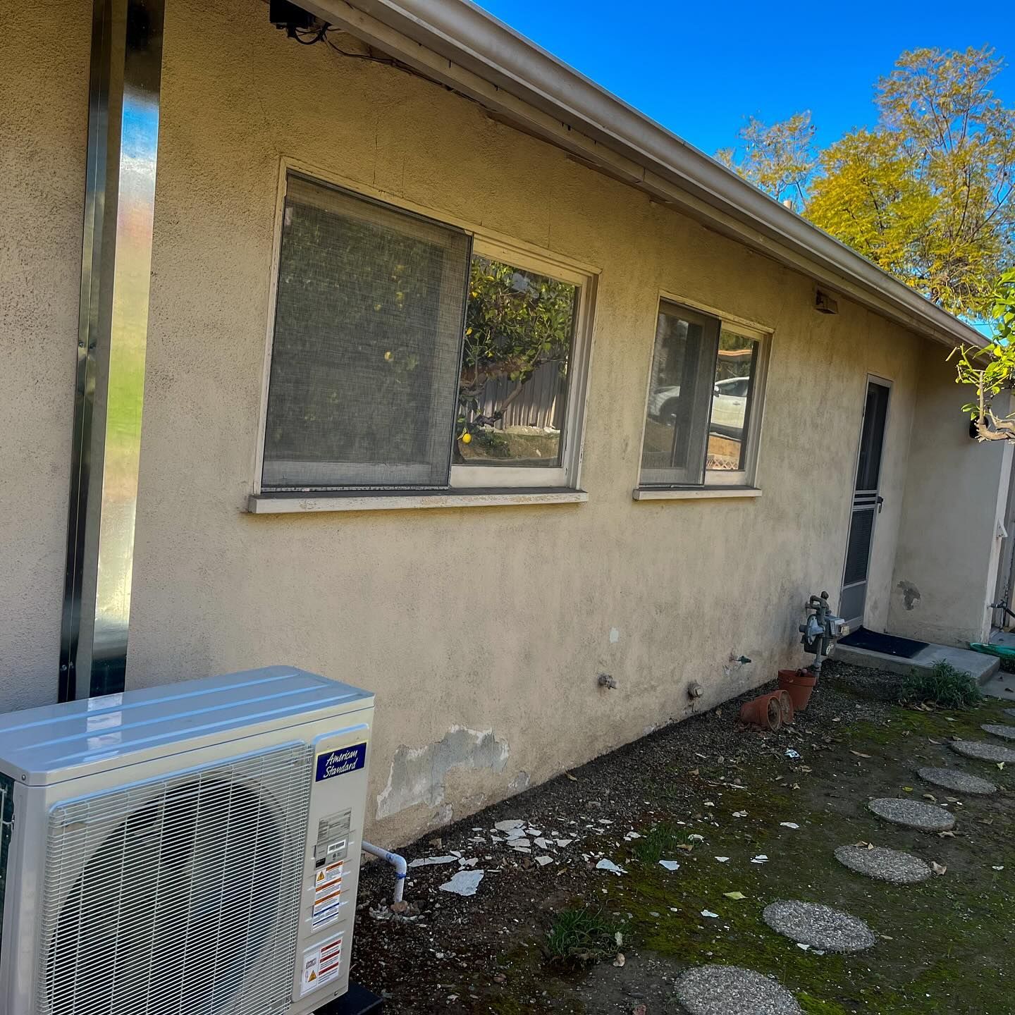 Side of a beige house with windows, a small path, and an outdoor air conditioner unit.