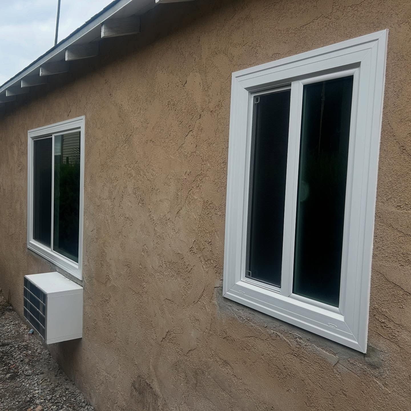 Exterior beige stucco wall with two white-framed windows and a small wall-mounted unit below one window