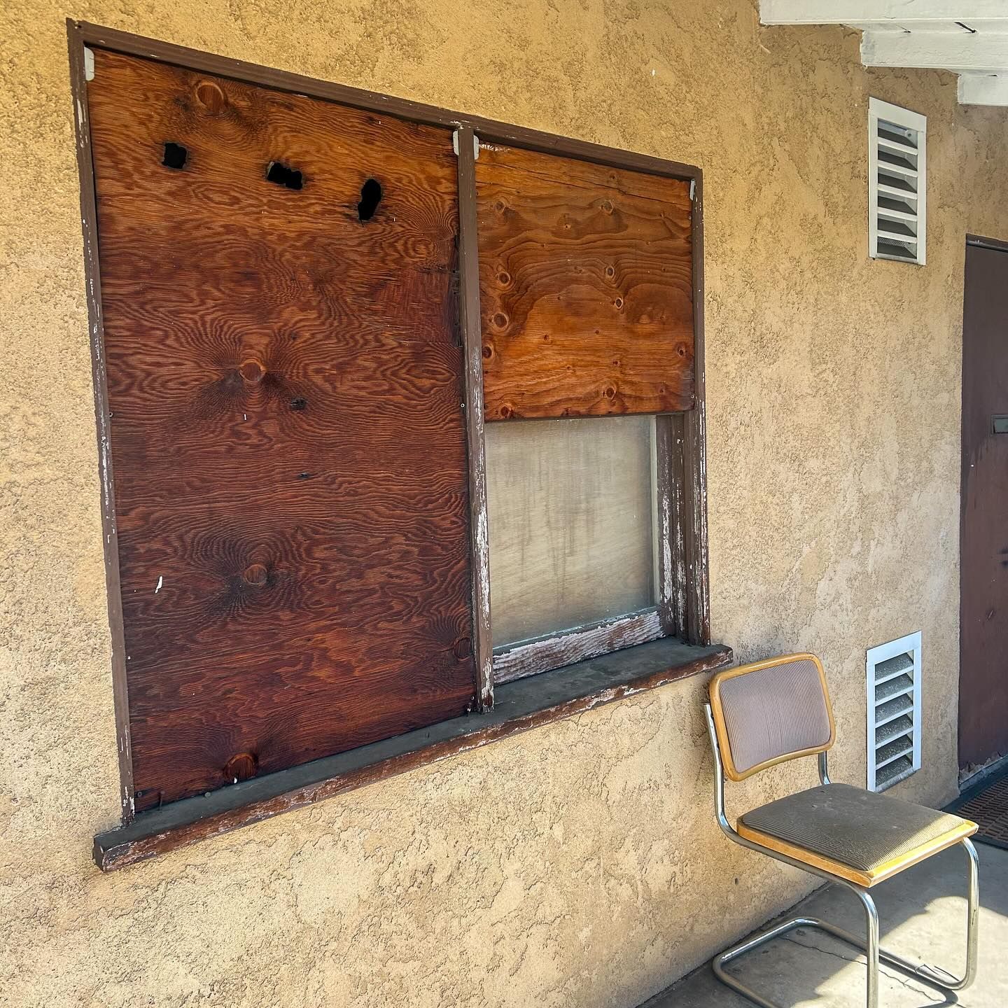 Closed wooden window on a stucco wall beside a chair and vent in an outdoor patio area