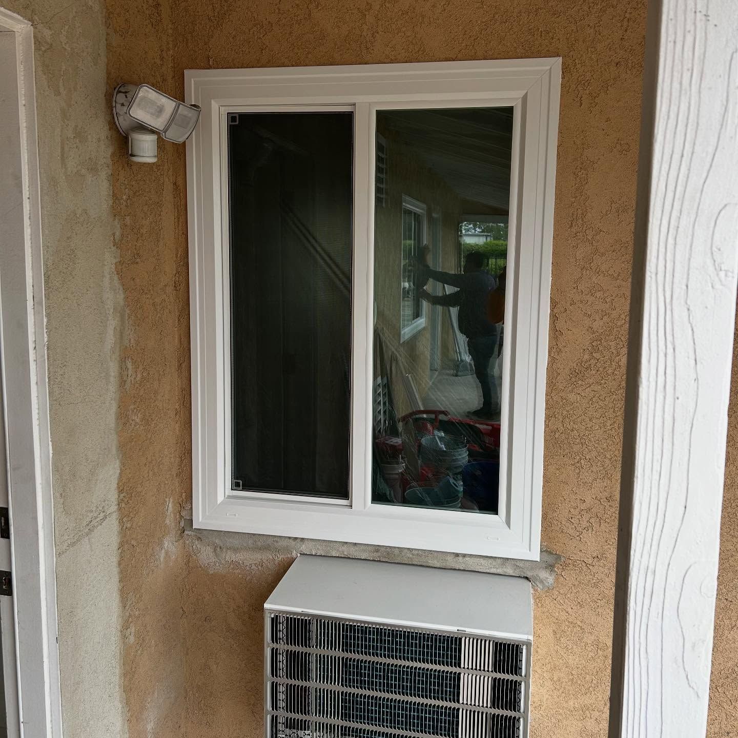 White-framed window above an outdoor HVAC unit on a stucco wall, with a security light nearby