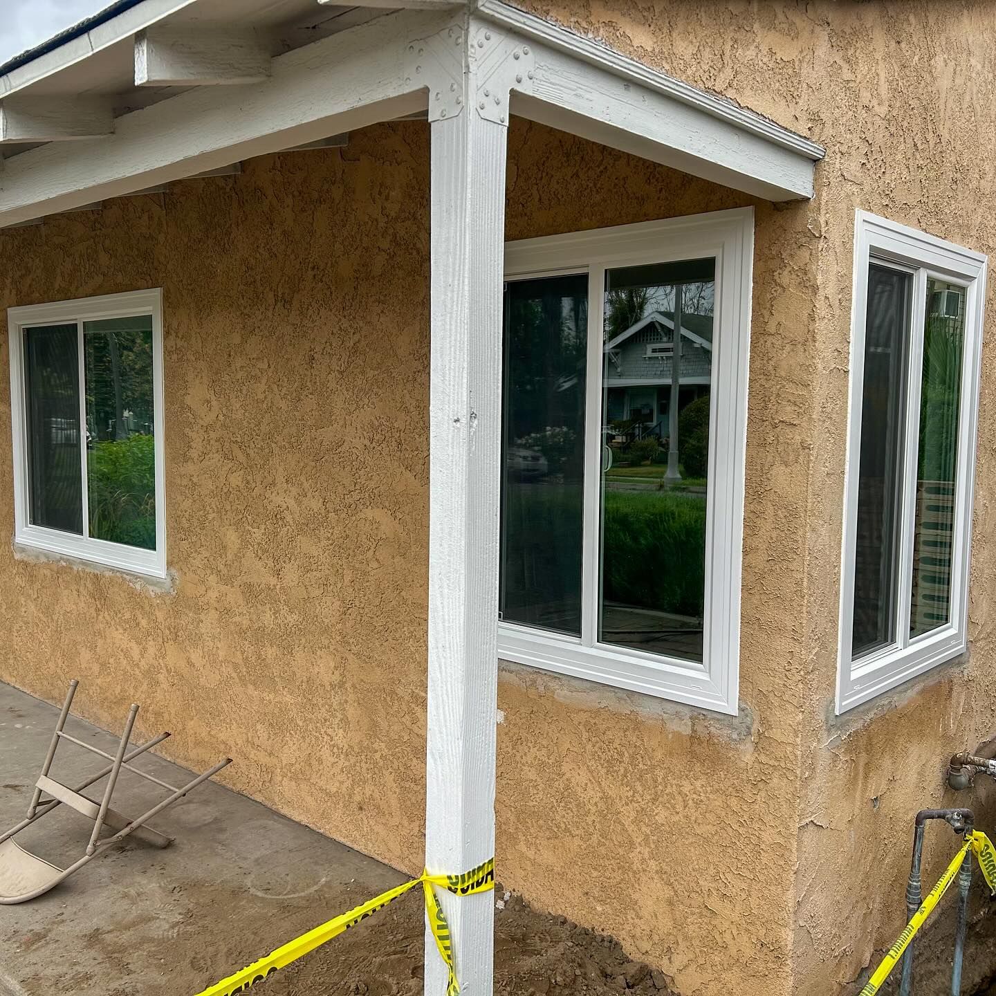 Partially renovated house exterior with tan stucco walls, white trim, and taped-off windows under a covered porch