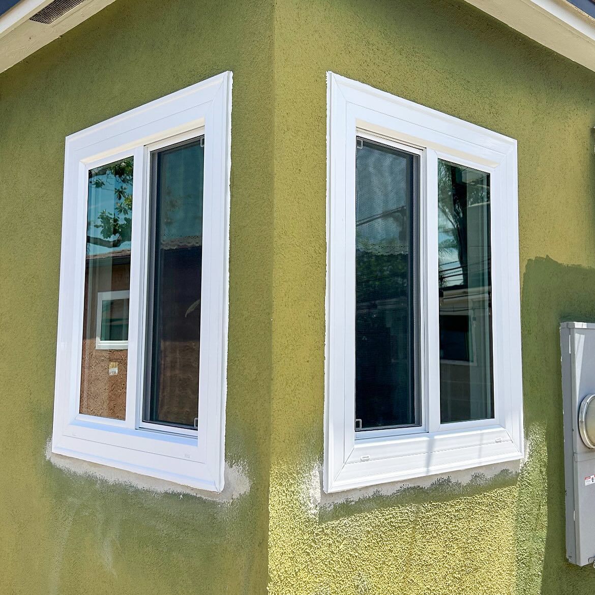 Corner of a green stucco house with two white-framed windows and a utility box on the right