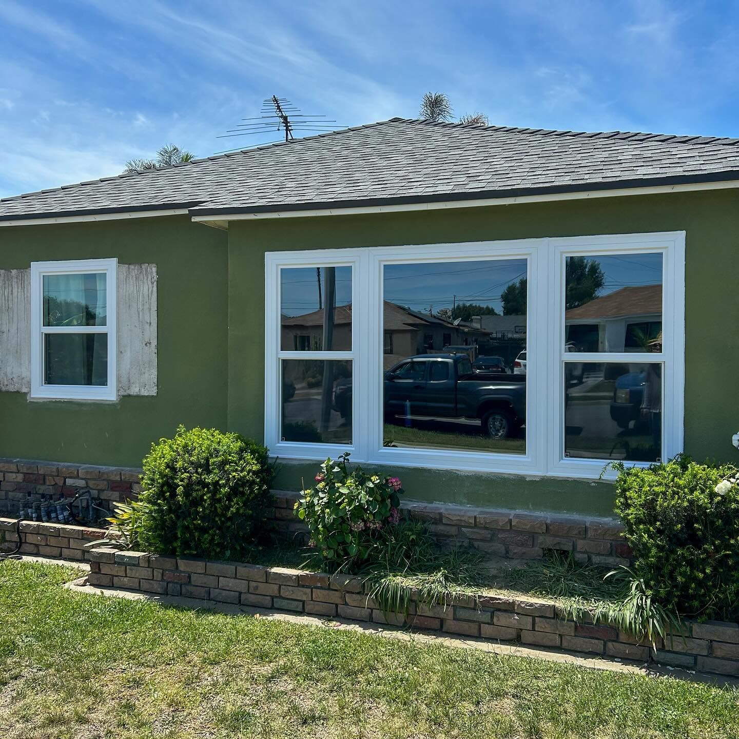 Single-story green house with white-trimmed windows, front shrubs, and a sunny lawn