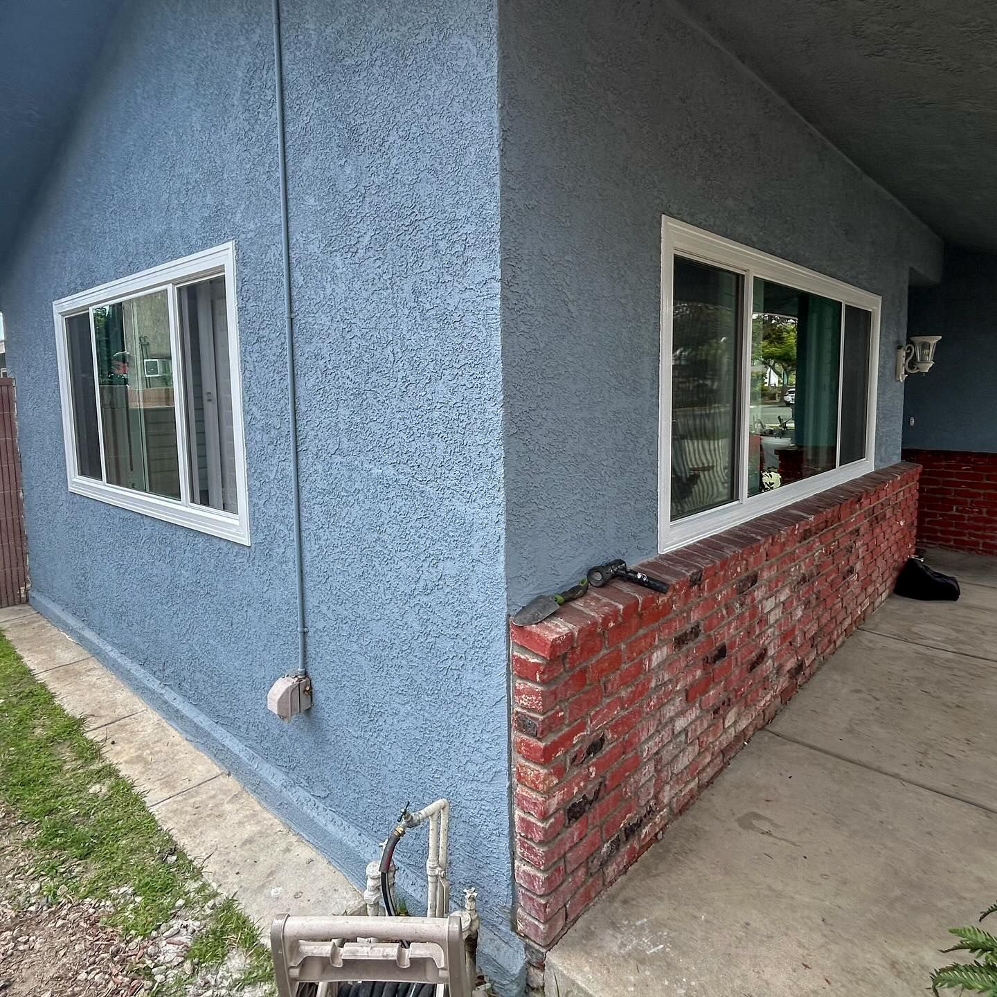 Blue stucco house corner with white-trimmed windows and a red brick base beside a concrete patio