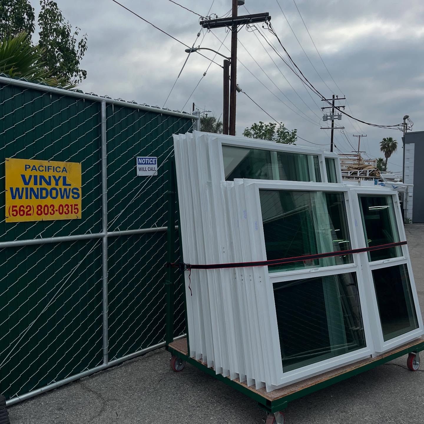 Stacked white vinyl windows on a cart beside a chain-link fence and sign in an industrial yard.