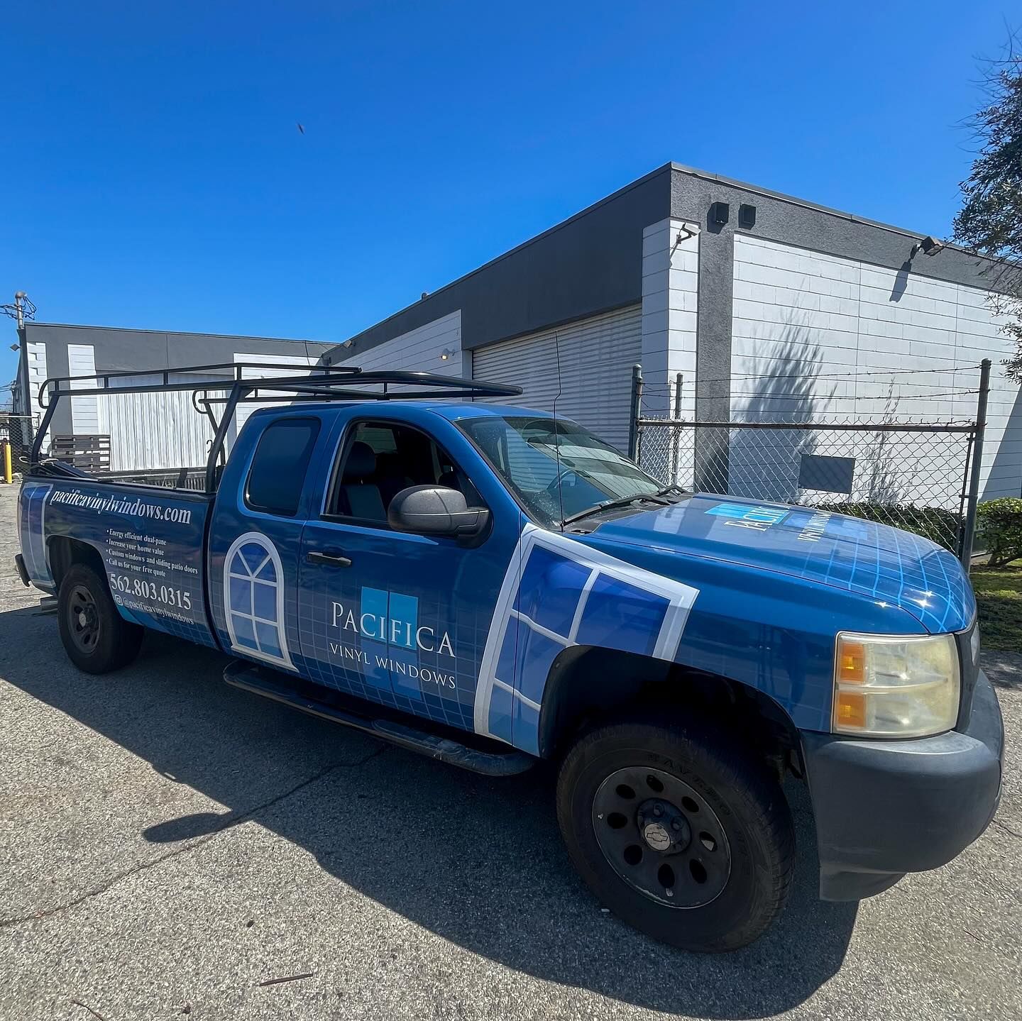 Blue utility pickup truck with ladder rack and company lettering parked beside a white building