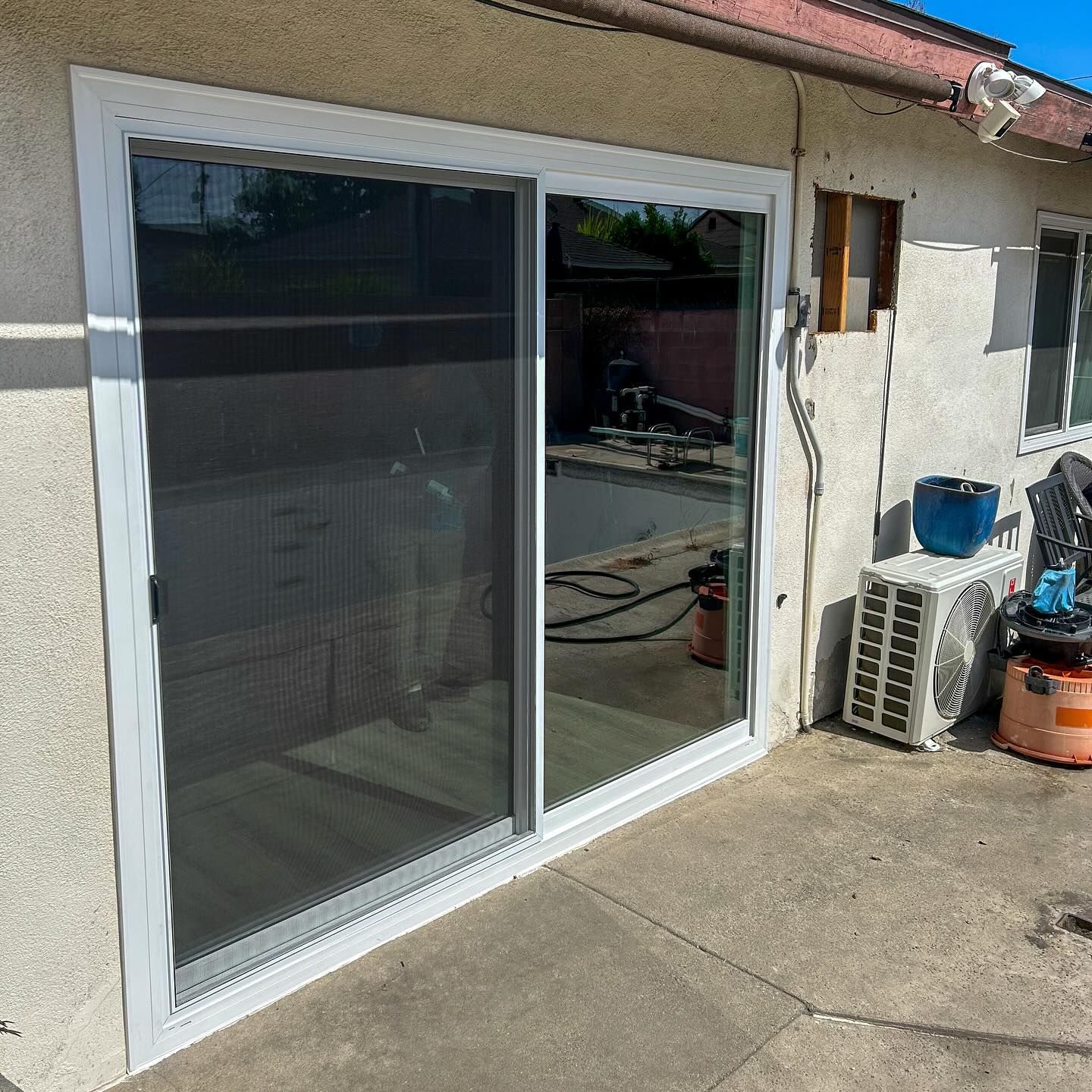 Sliding glass doors with white trim on a stucco exterior wall, next to an air conditioner unit and patio items.