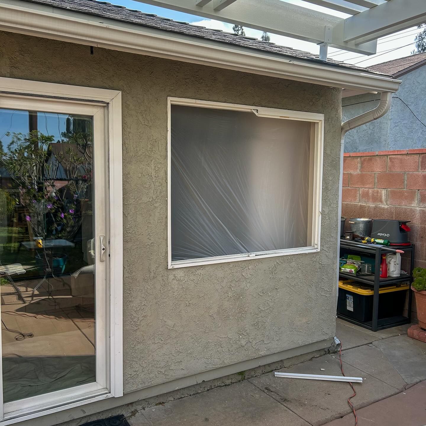 Patio beside a house with a sliding glass door, screened window, and storage shelves near a brick wall.