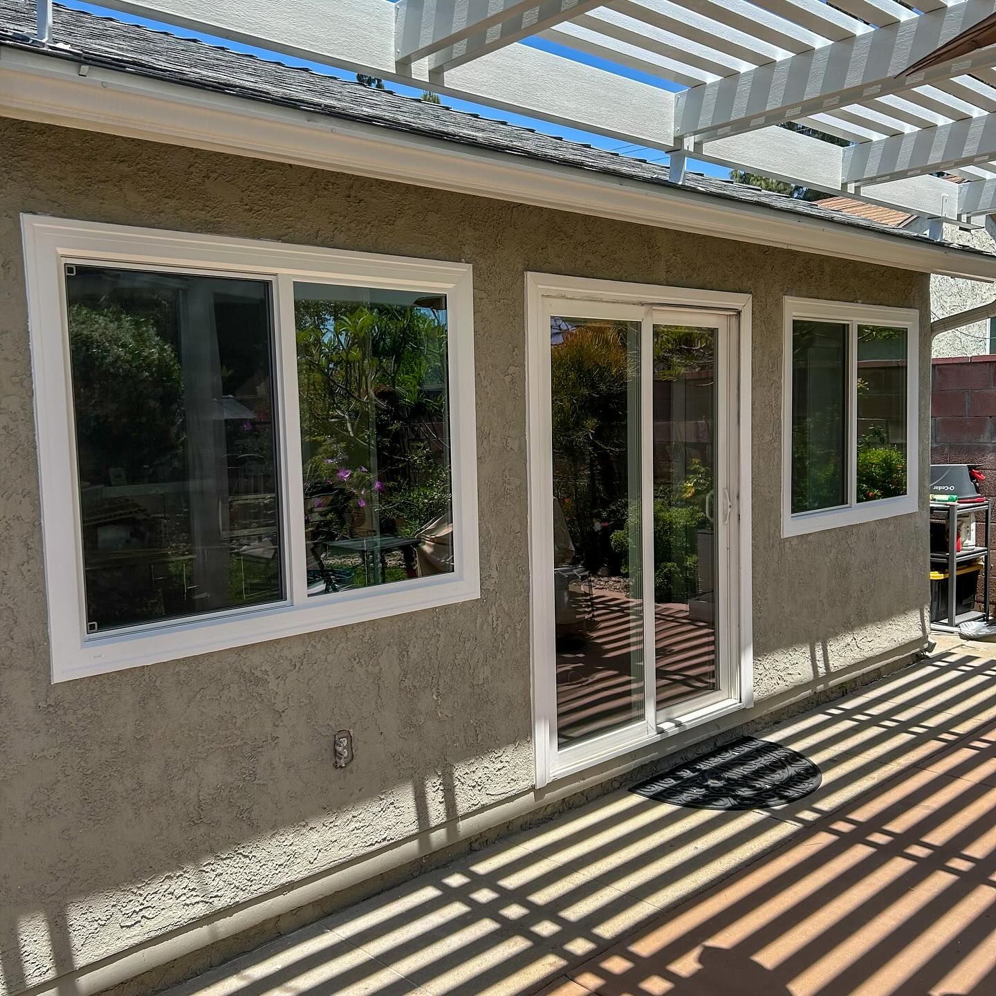 Stucco house exterior with white-framed windows and glass door under a slatted patio roof, casting striped shadows