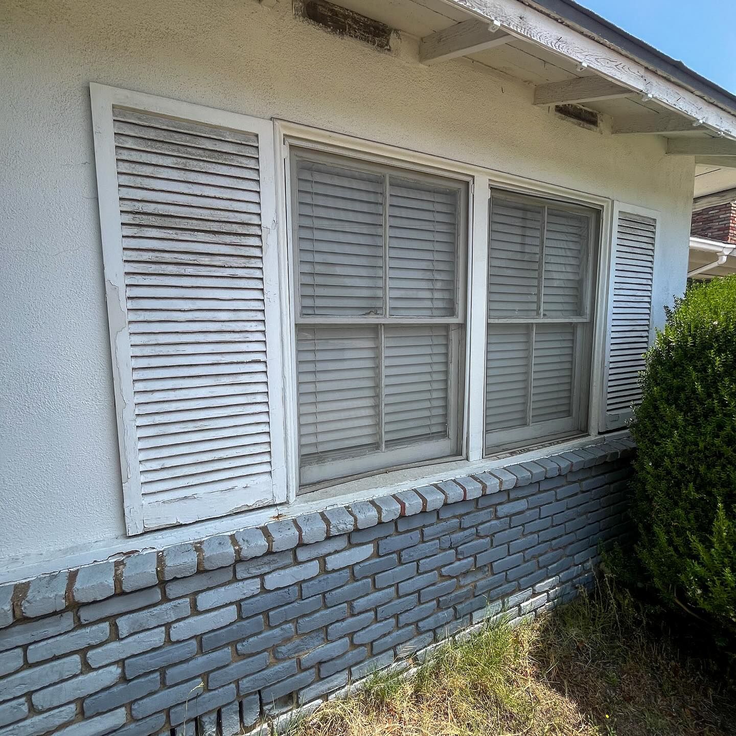 White house exterior with shuttered windows, gray brick base, and a green shrub in front