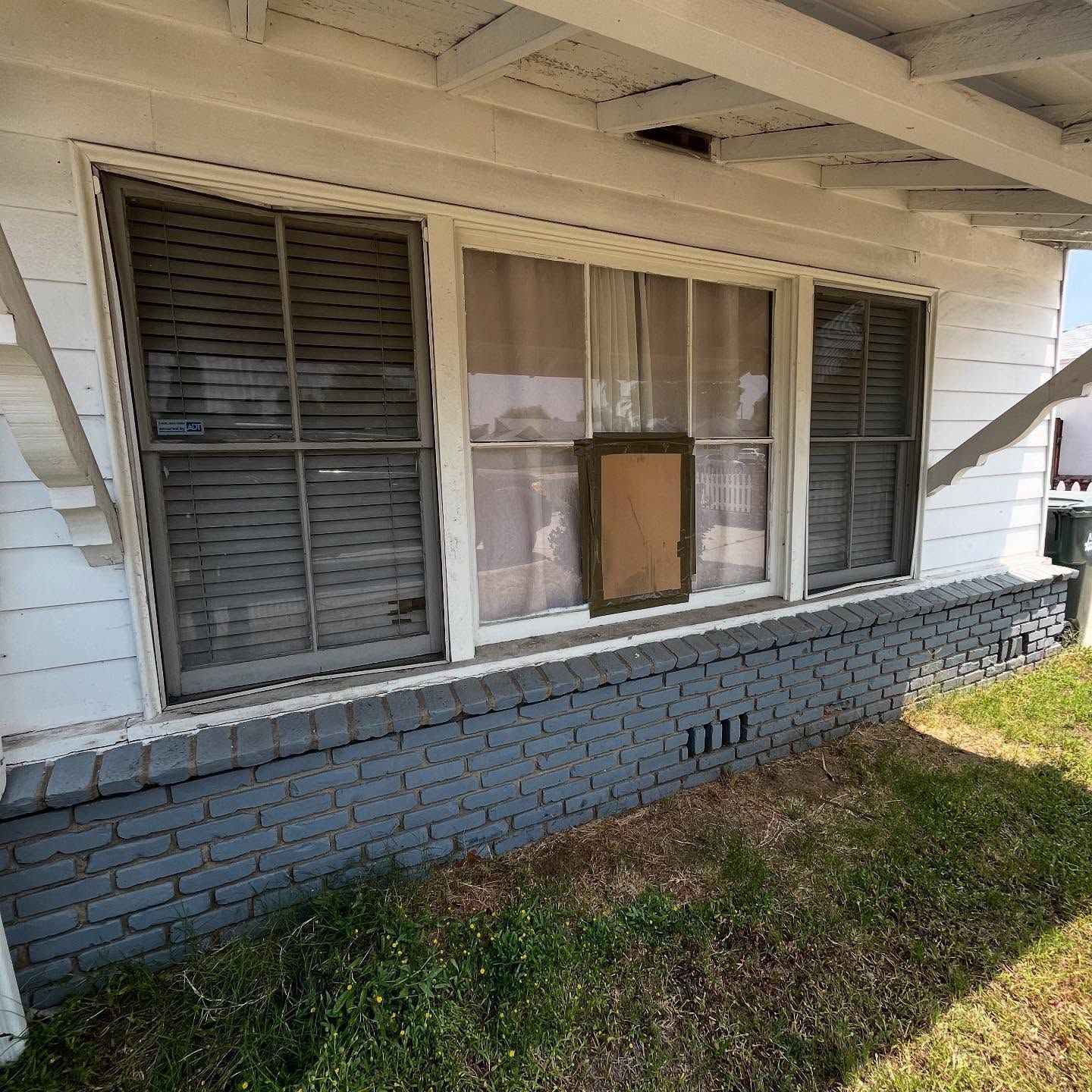 Weathered house exterior with boarded window, gray shutters, blue brick foundation, and grassy yard