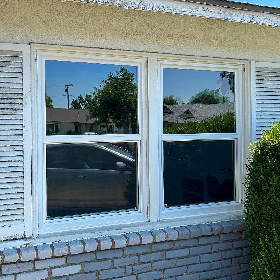 Exterior window with white shutters reflecting trees and a parked car on a brick house wall