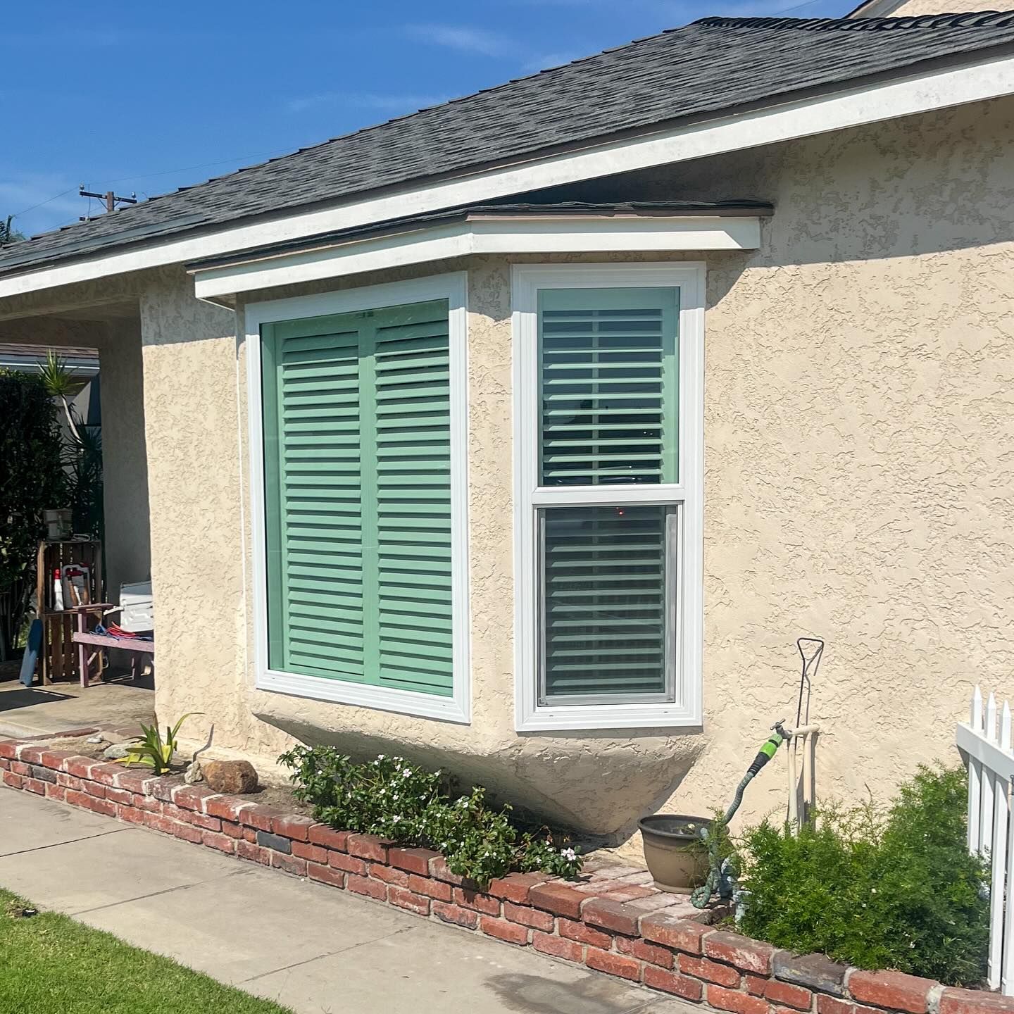 Beige stucco house exterior with green shutters and a small front garden beside a sidewalk