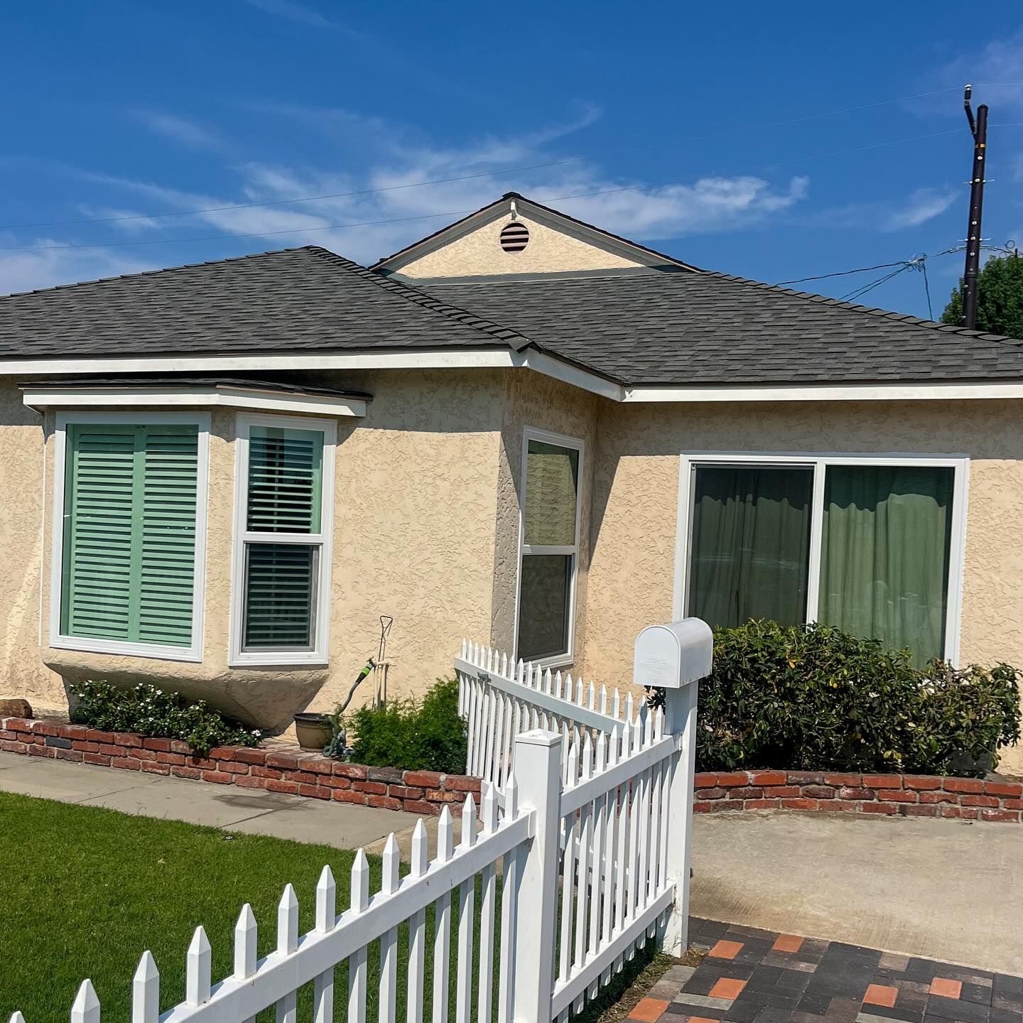 Single-story beige house with white fence, green shutters, and a small front yard under a blue sky
