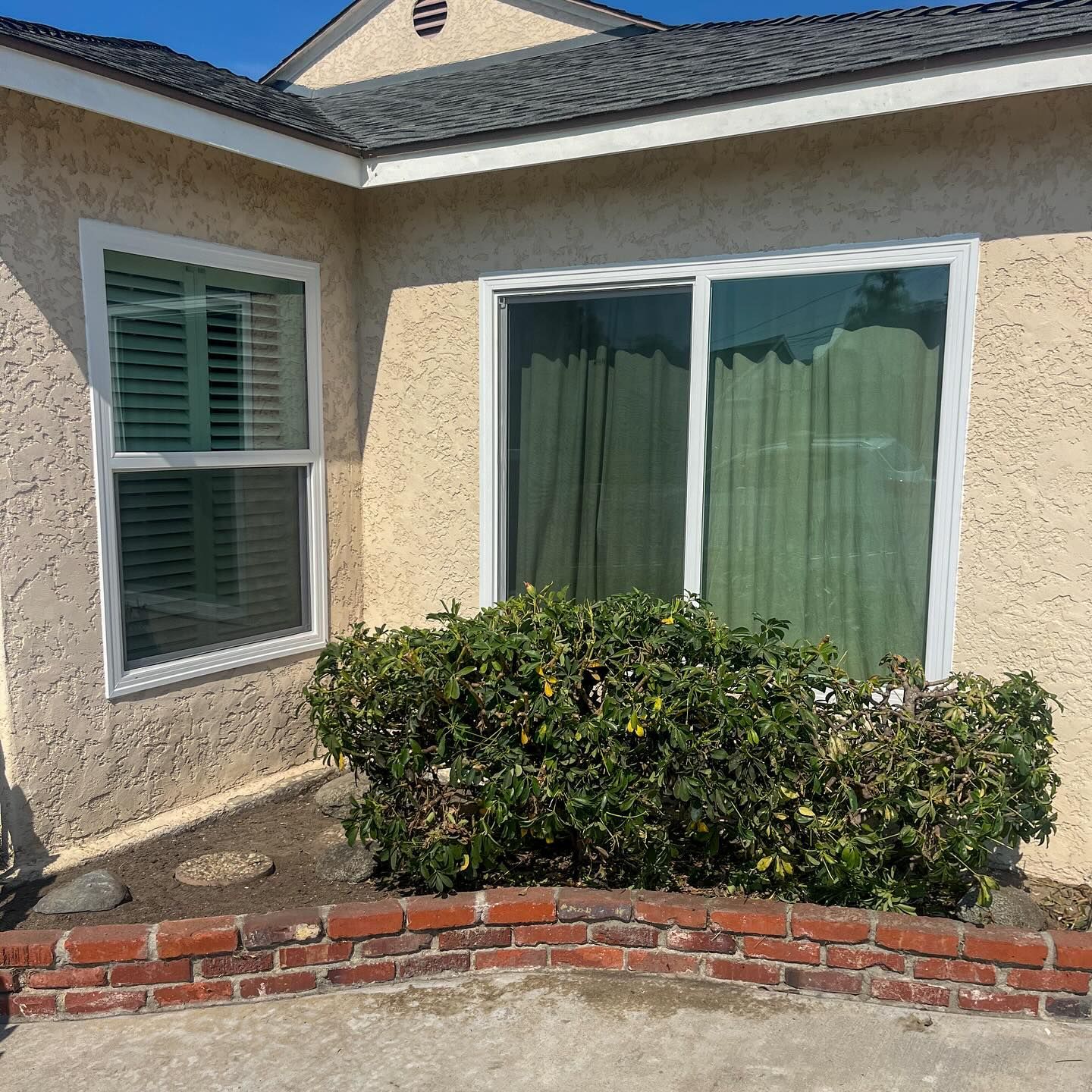Stucco house exterior with white-trimmed windows, sliding glass door, shrubs, and a low brick planter wall.