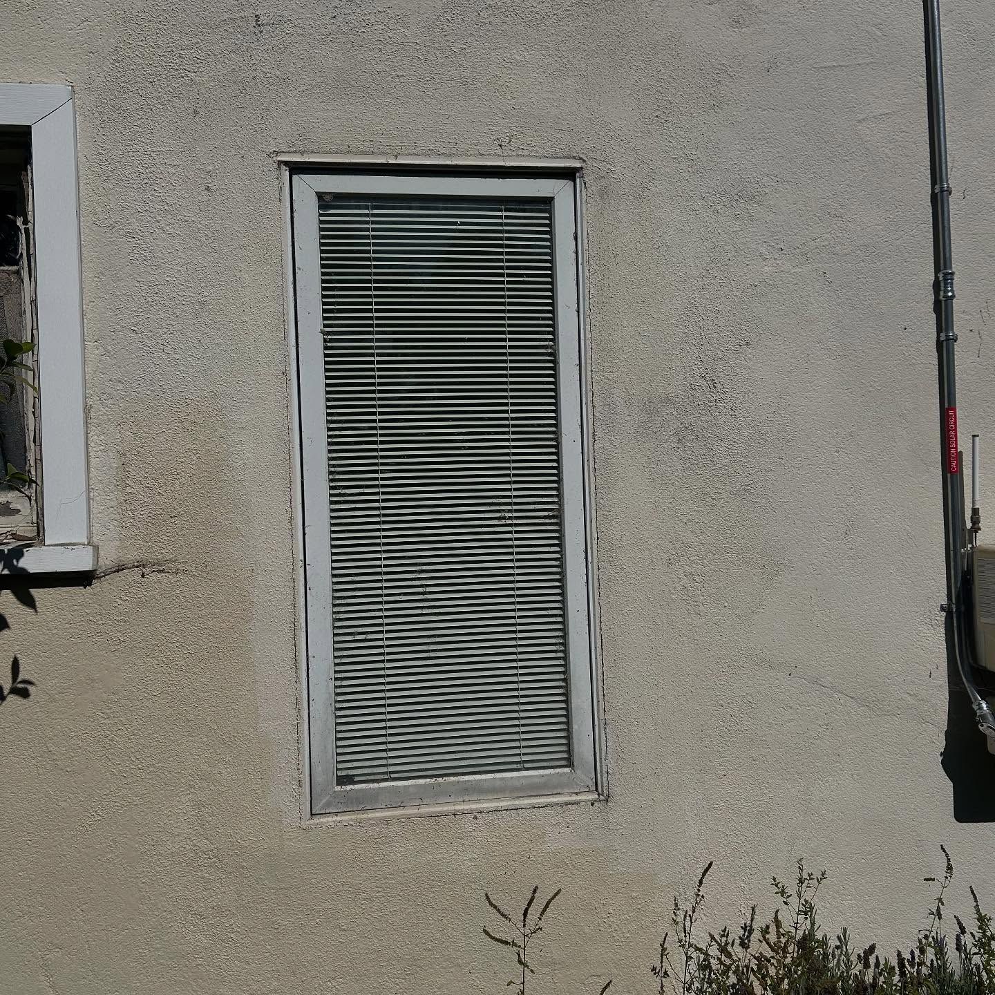 Narrow window with white blinds set in a beige stucco exterior wall.