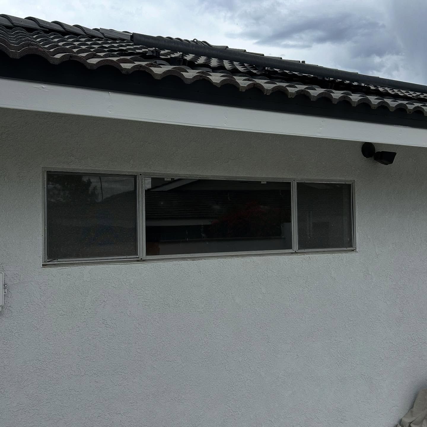 White stucco house wall with a narrow horizontal window and dark tiled roof under cloudy sky