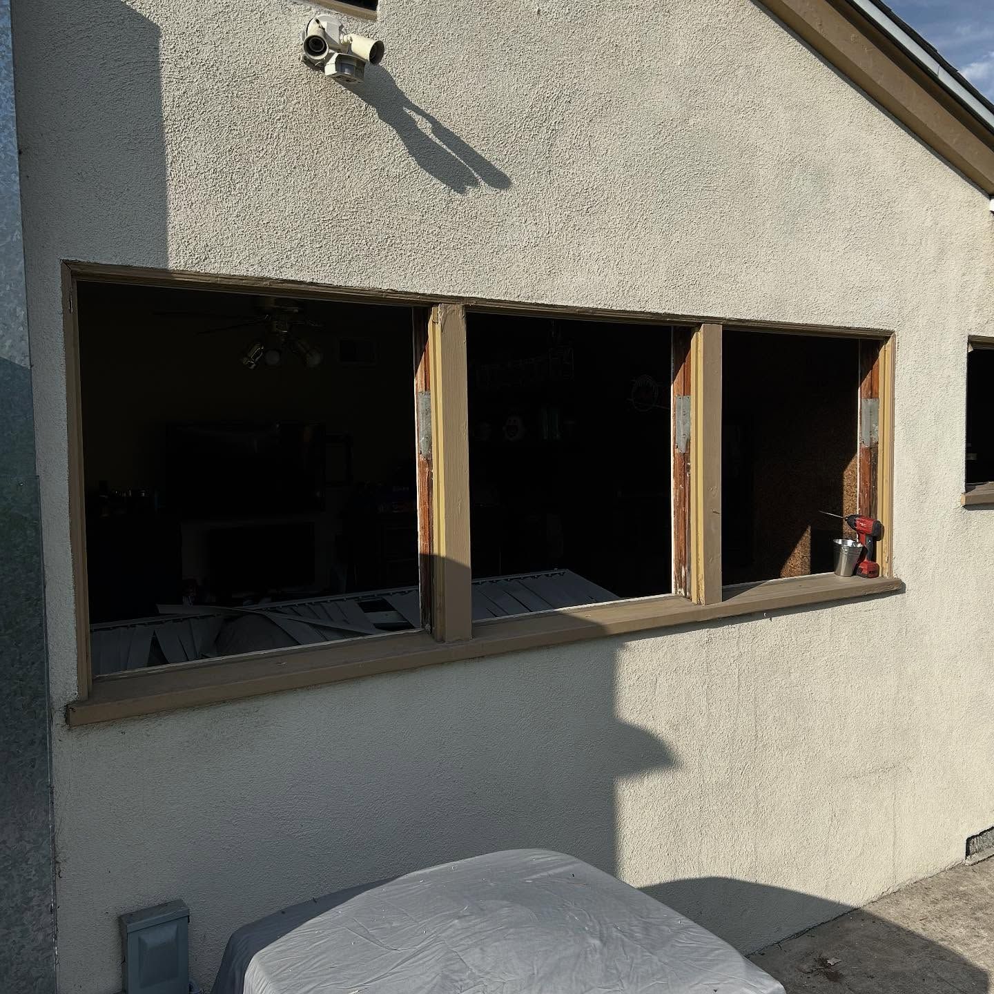 House exterior with three open window frames and a light gray wall, sunlight casting shadows.