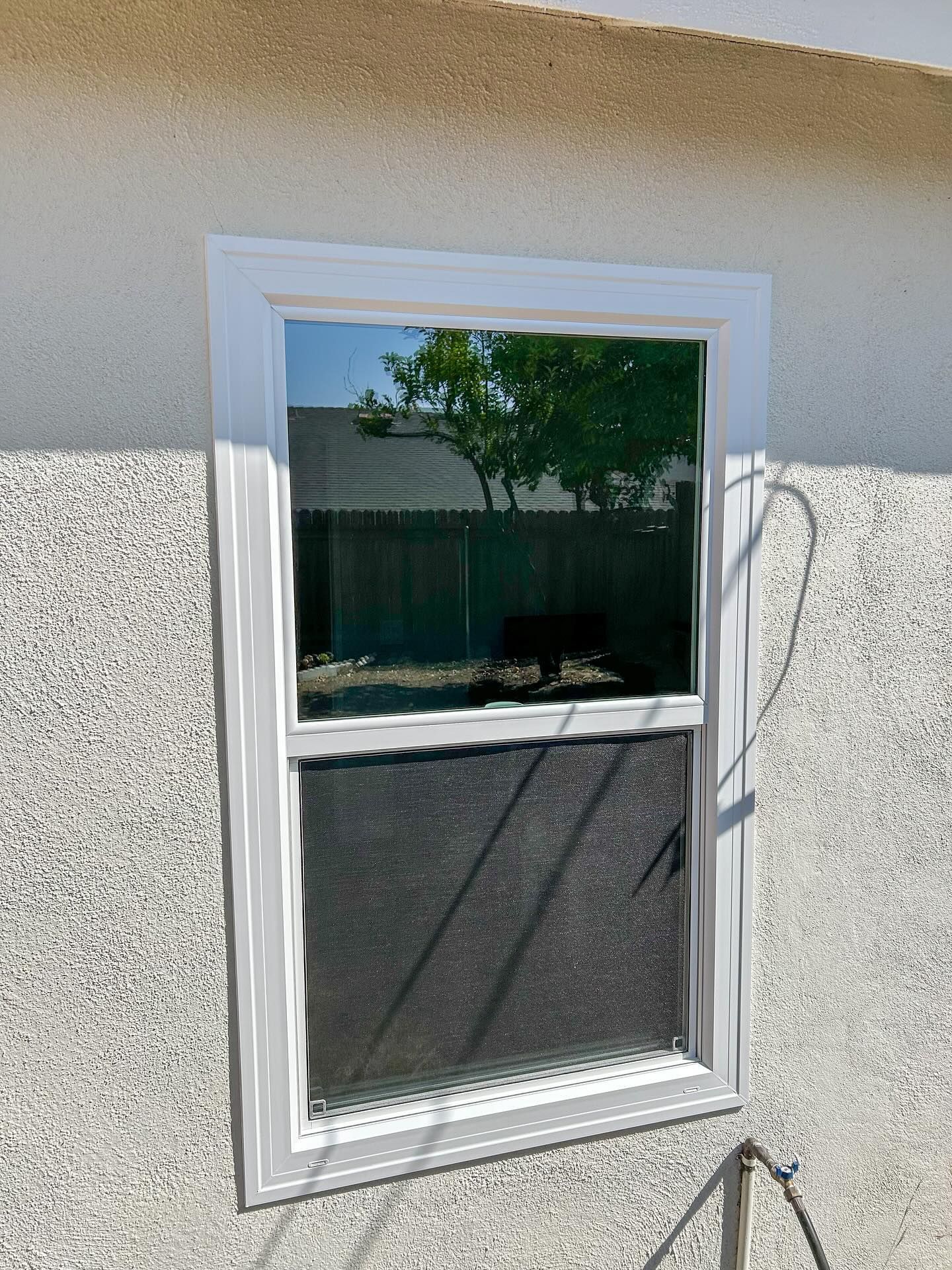 White-framed window with a dark screen on a beige exterior wall, with a reflection of trees.