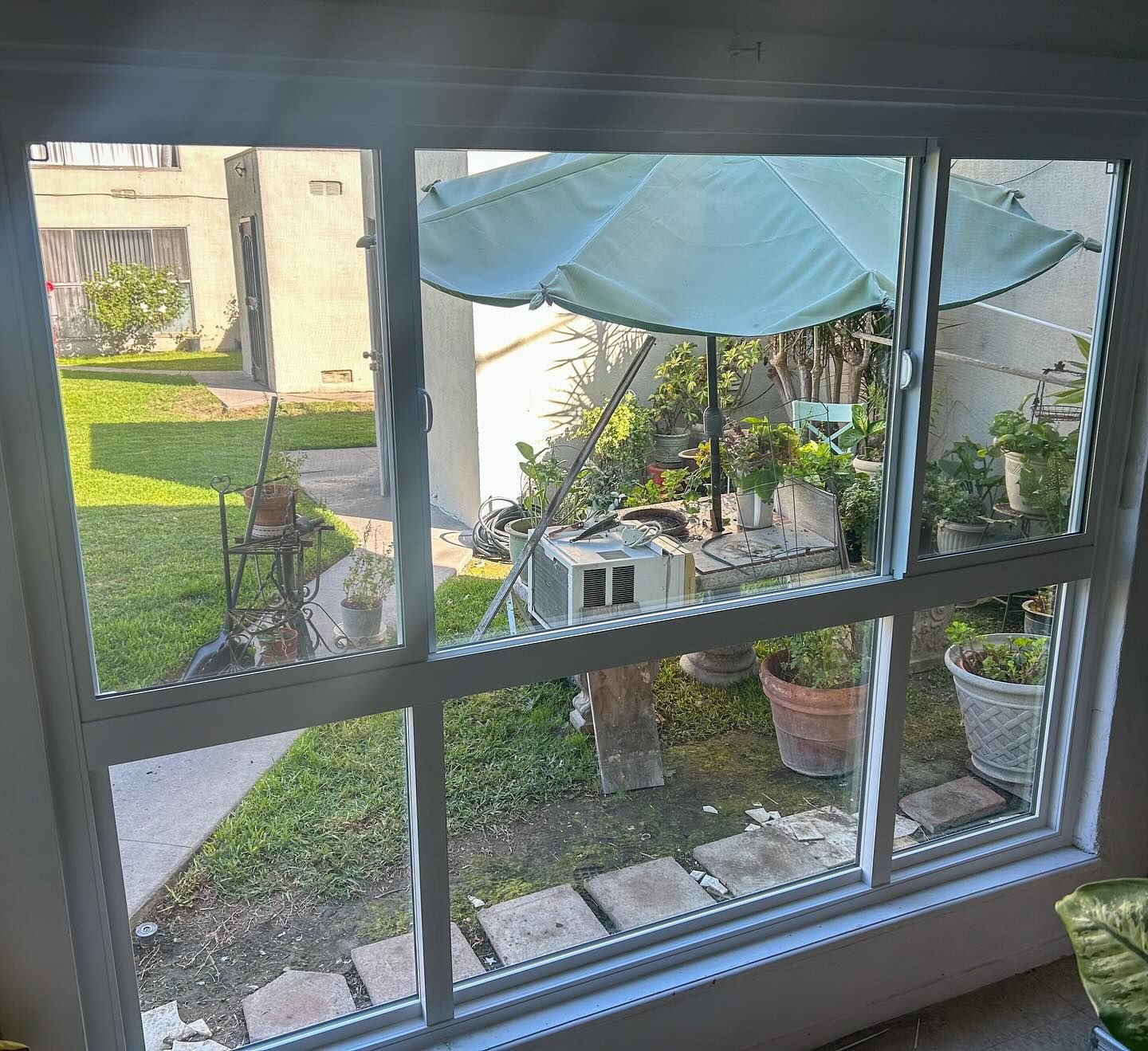 View through a window to a small courtyard with patio table, potted plants, and blue shade sail