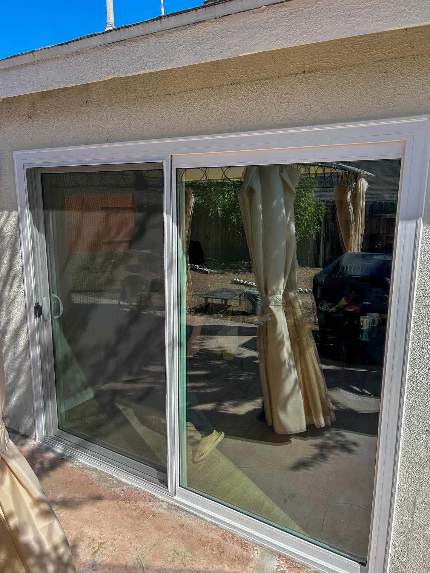 Sliding glass patio door with curtains, reflecting a backyard and tree shadows.