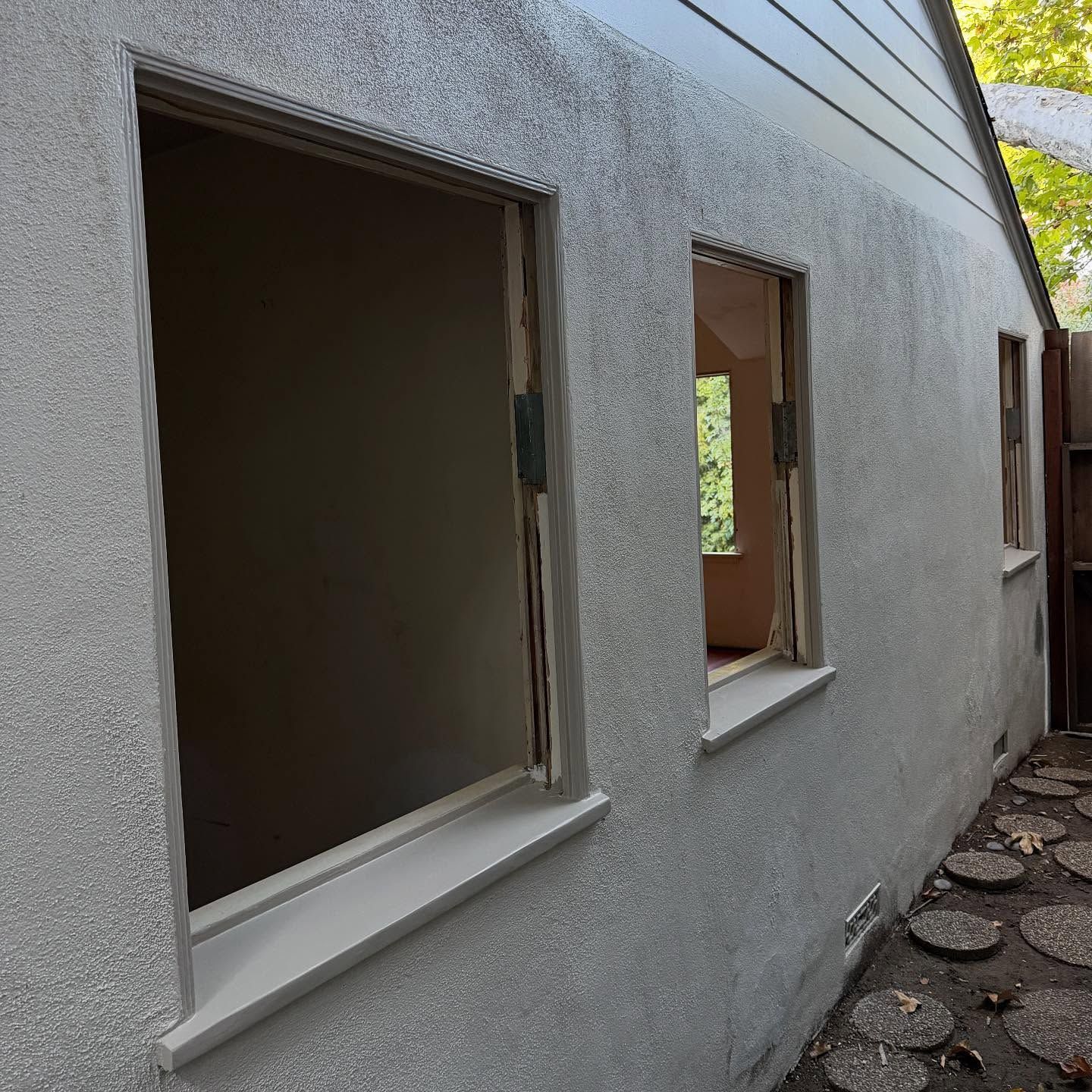 Stucco exterior wall with three open window openings along a narrow stone path