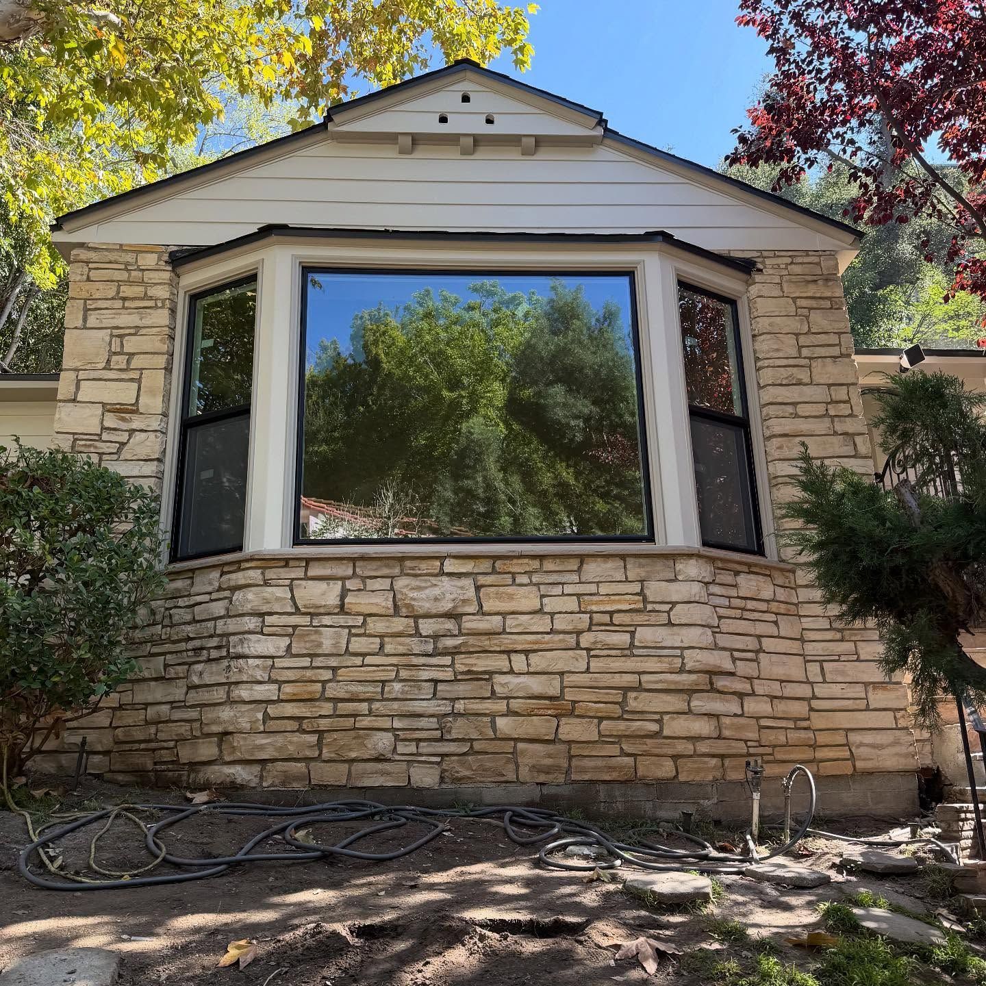 Stone-front house facade with a large bay window and a small gabled roof, surrounded by trees and yard debris