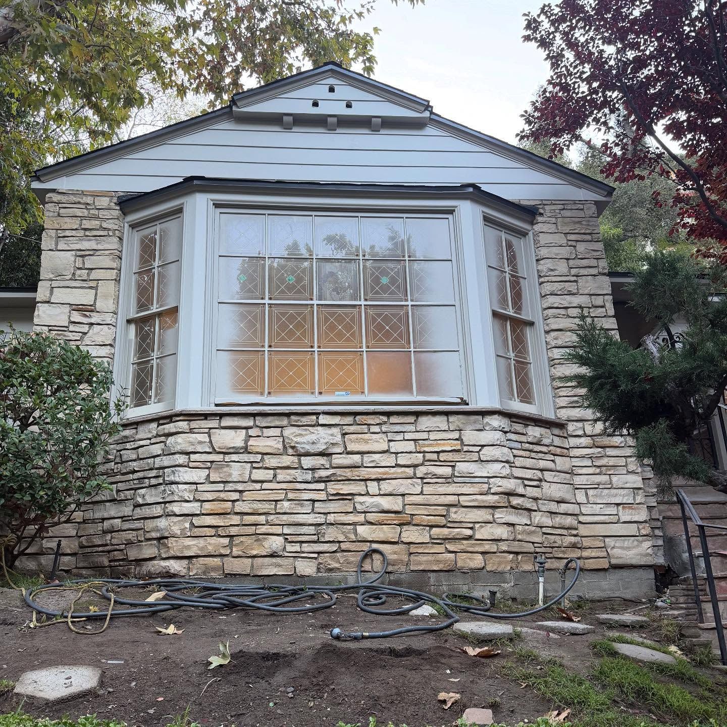 Stone house front with a bay window, white trim, and a small gabled roof, surrounded by trees and yard.