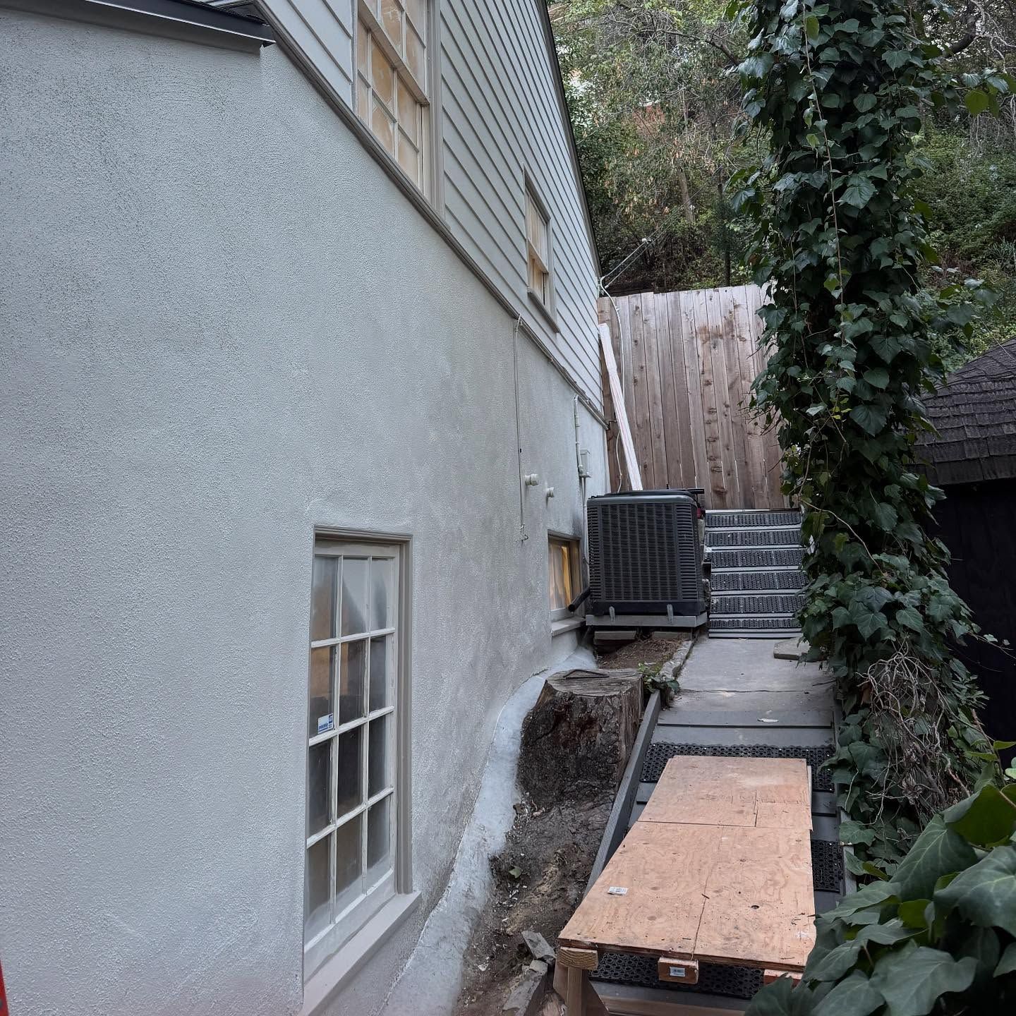 Narrow side yard beside a gray house, with steps, an air conditioner unit, and plywood covering the ground.
