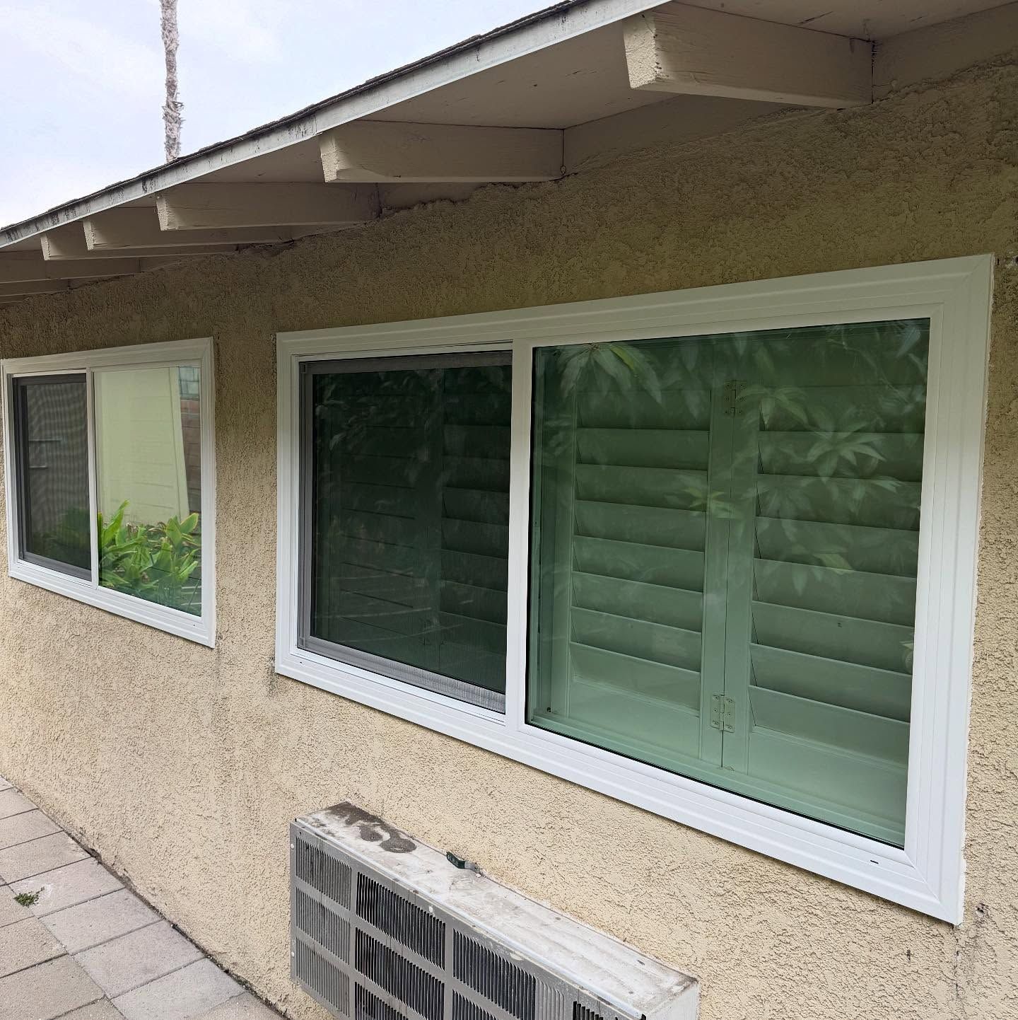 Exterior wall of a stucco house with two windows, green shutters, and an air conditioner unit below.