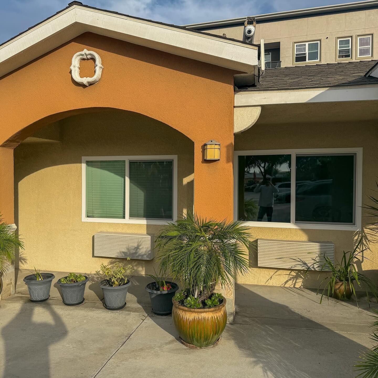 Front of a tan stucco house with arched entry, two windows, potted plants, and a sunlit concrete patio