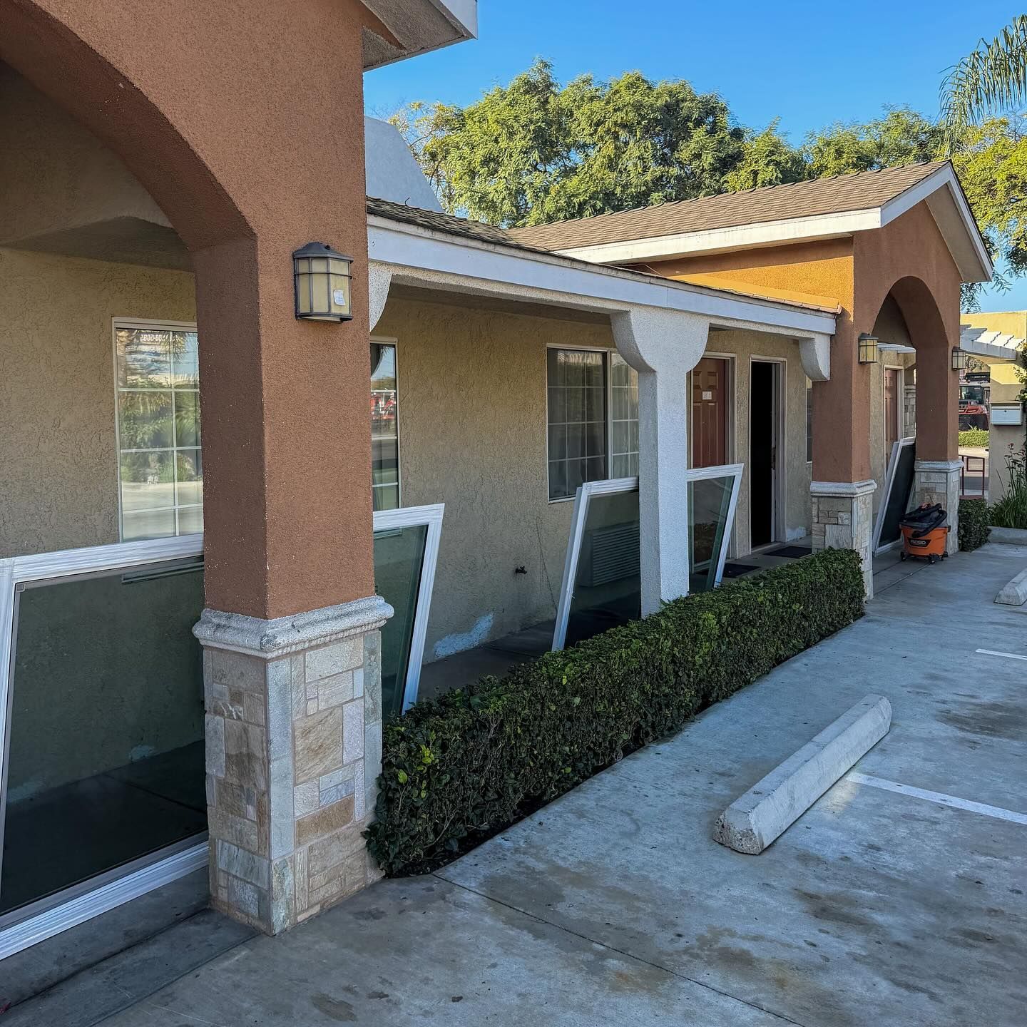 Exterior walkway of a beige stucco building with white columns, large windows, and hedges along a concrete path