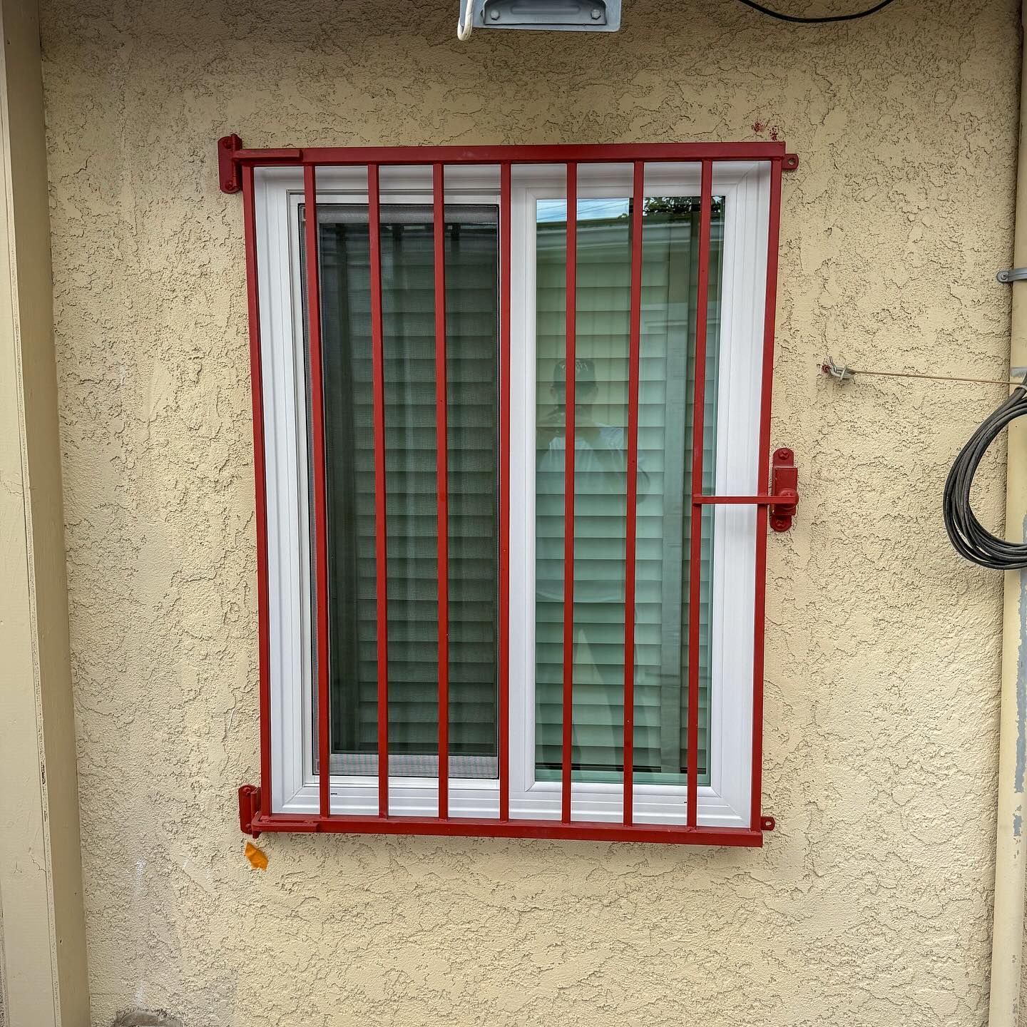 Red barred window with white curtains and green blinds on a beige stucco wall.