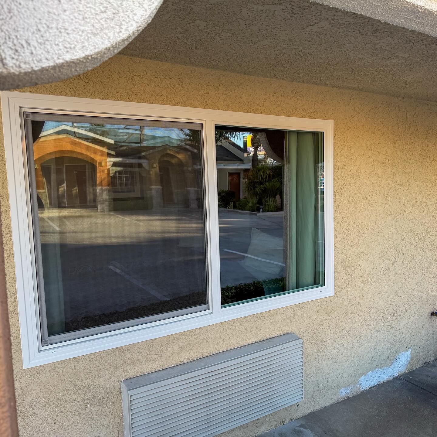 White-framed window set in a beige stucco wall, with a vent below and street reflections in the glass